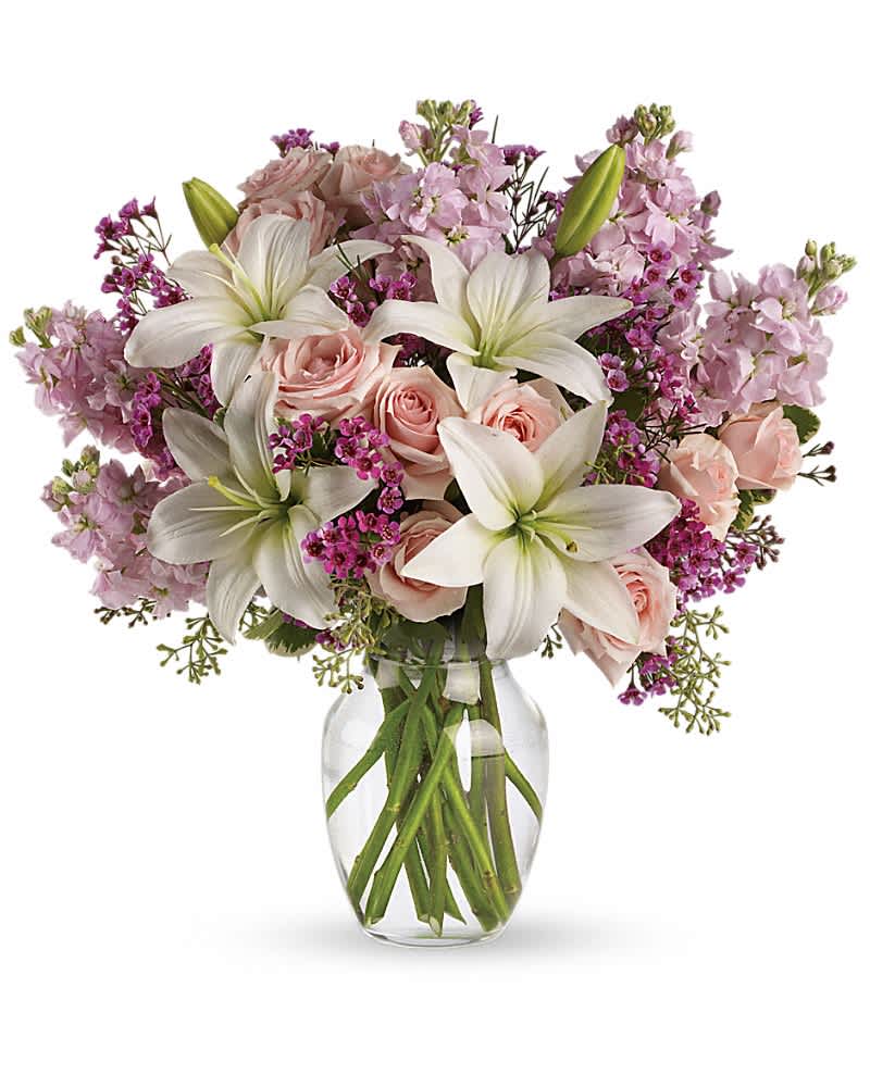 Pastel bouquet of white lilies, pink roses, and lavender flowers arranged in a clear glass vase.