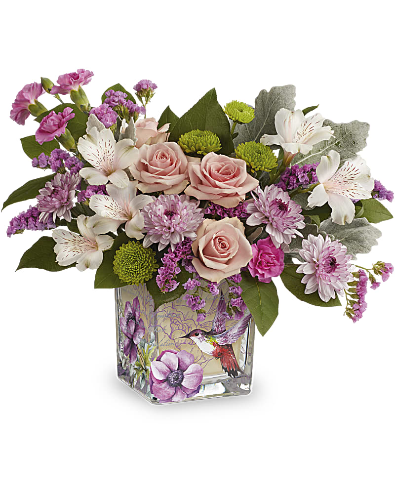 Short arrangement of pink roses, white lilies, and purple blooms in a glass cube vase with hummingbird art.