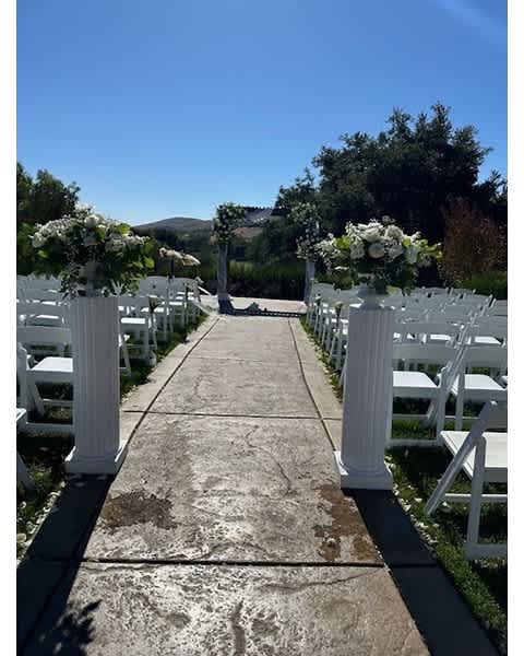 Outdoor wedding aisle with white flower arrangements on tall white columns and an arch in the distance