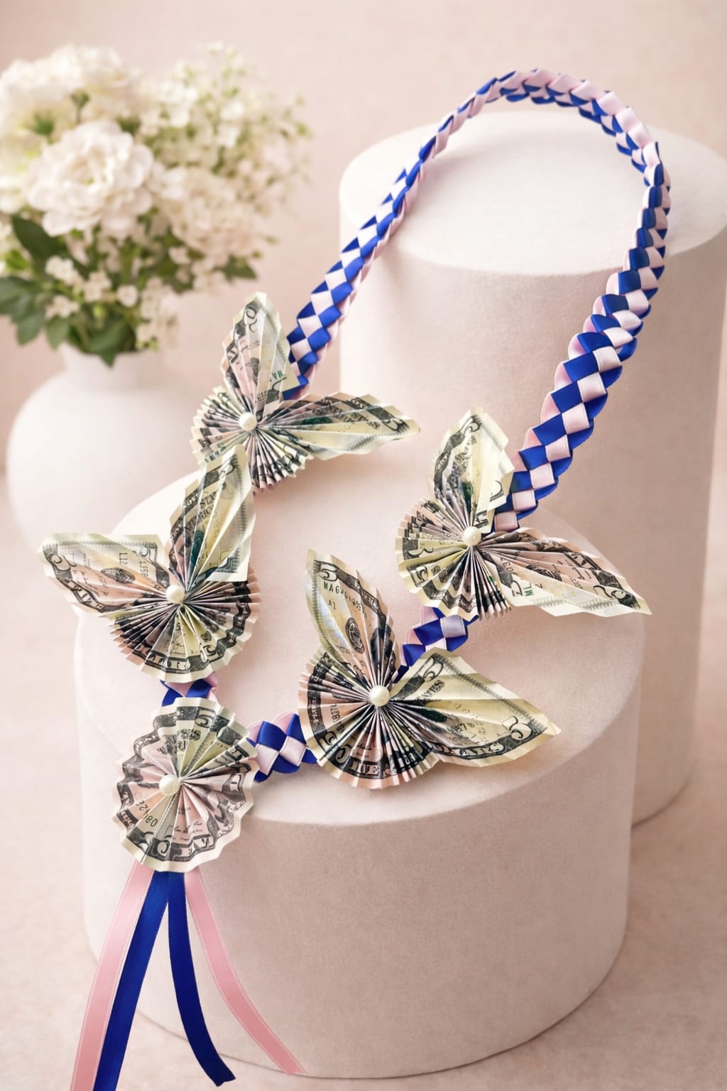 Lei of folded dollar butterflies - Celebrate graduation with a meaningful and eye-catching gift that combines beauty and value. This handcrafted butterfly money lei features intricately folded currency arranged into delicate butterfly shapes, accented with pearl centers for an elegant touch.  The lei is finished with a premium braided ribbon design in soft pink and royal blue tones, creating a modern and stylish look that stands out in photos and ceremonies.  Perfect for graduation, birthdays, or special achievements, this lei is both a keepsake and a gift — allowing the recipient to enjoy the design and later unfold the bills.  Details: • Handmade butterfly money folds with pearl accents • Braided ribbon in pink &amp; blue color combination • Lightweight and comfortable to wear • Each piece is uniquely crafted  Perfect For: • Graduation gifts  • Celebration moments • Photo-ready statement piece  Note: • Bill denominations can be customized upon request • Colors can be adjusted to match school theme • Due to handmade nature, slight variations may occur