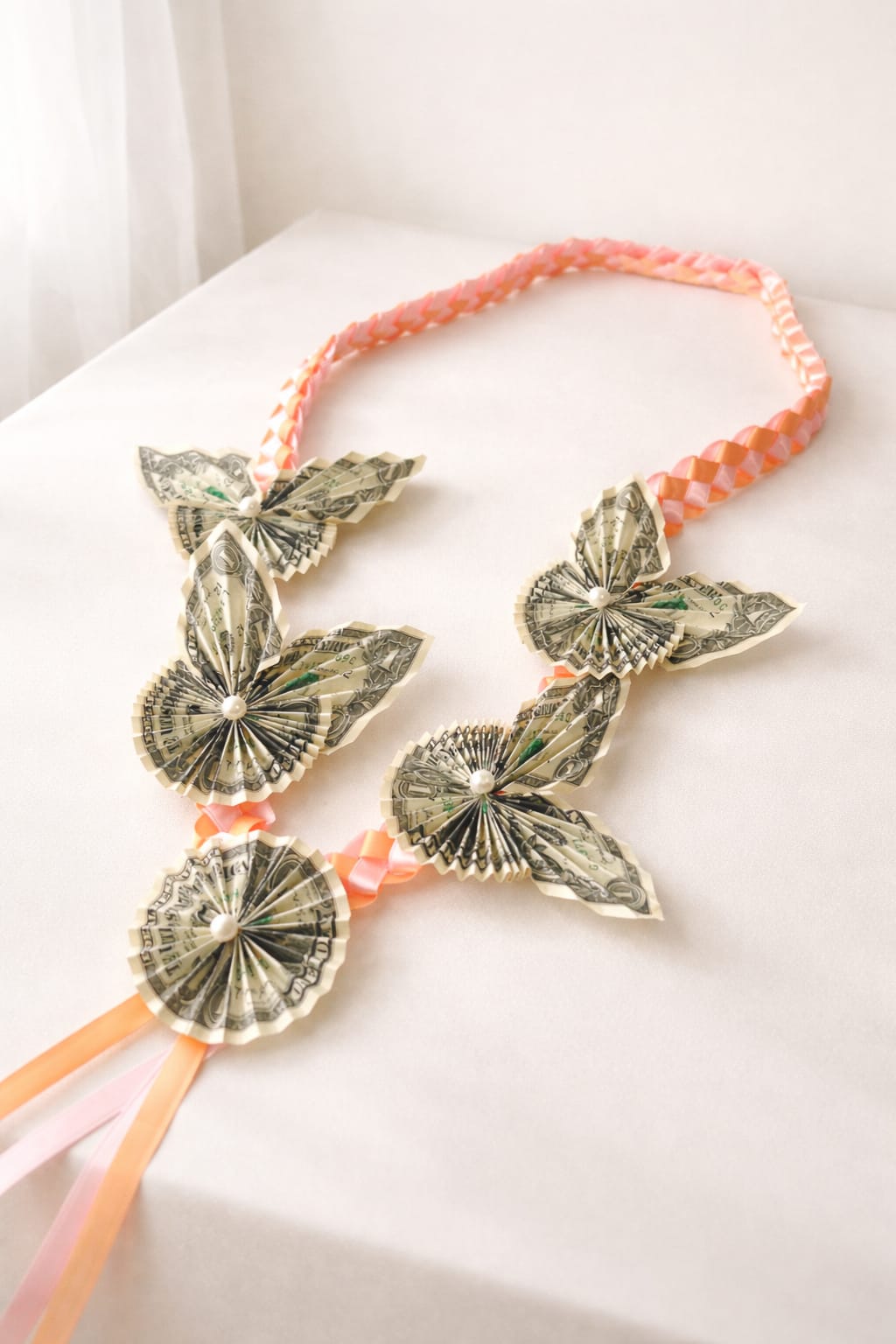 Money lei with delicate dollar flowers - Celebrate a meaningful milestone with our handcrafted Butterfly Blossom Money Lei, a unique and eye-catching graduation gift designed to impress. Each lei features delicately folded dollar bills shaped into elegant butterflies and rosette blooms, symbolizing growth, transformation, and success.  Accented with soft peach and blush satin ribbons, this lei brings a warm, joyful tone that perfectly complements graduation attire. The intricate hand-folded details and pearl-centered flowers add a refined, luxurious touch—making it not just a gift, but a memorable keepsake.  Perfect for graduations, celebrations, or special achievements, this money lei is a beautiful way to give both flowers and a meaningful monetary gift in one stunning design.