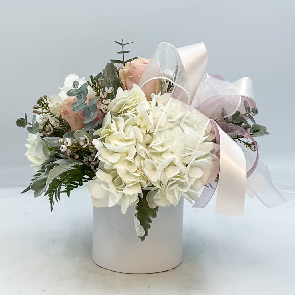 White hydrangeas and blush roses in a white cylinder vase with sheer white and pink ribbon bow