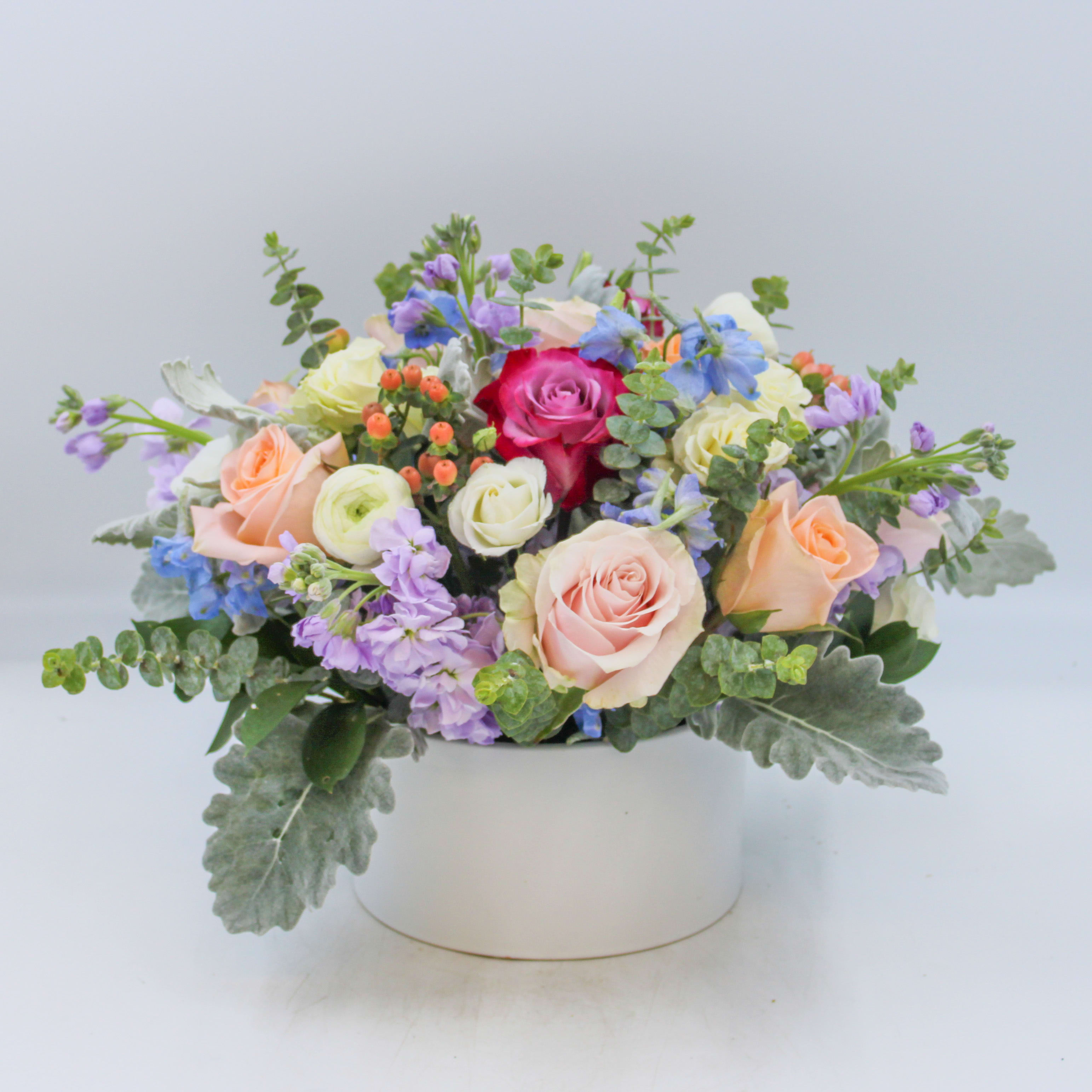 Pastel Grande - Pastel Grande is a colorful display of fresh florals arranged in a keepsake white ceramic dish. Pastel Grande includes Peach, Lavender and Blush Pink Roses, White Ranunculus, White Lisianthus, Peach Hypericum Berries, White Spray Roses, Blue Delphinium, Lavender Stock, Dusty Miller and Eucalyptus.