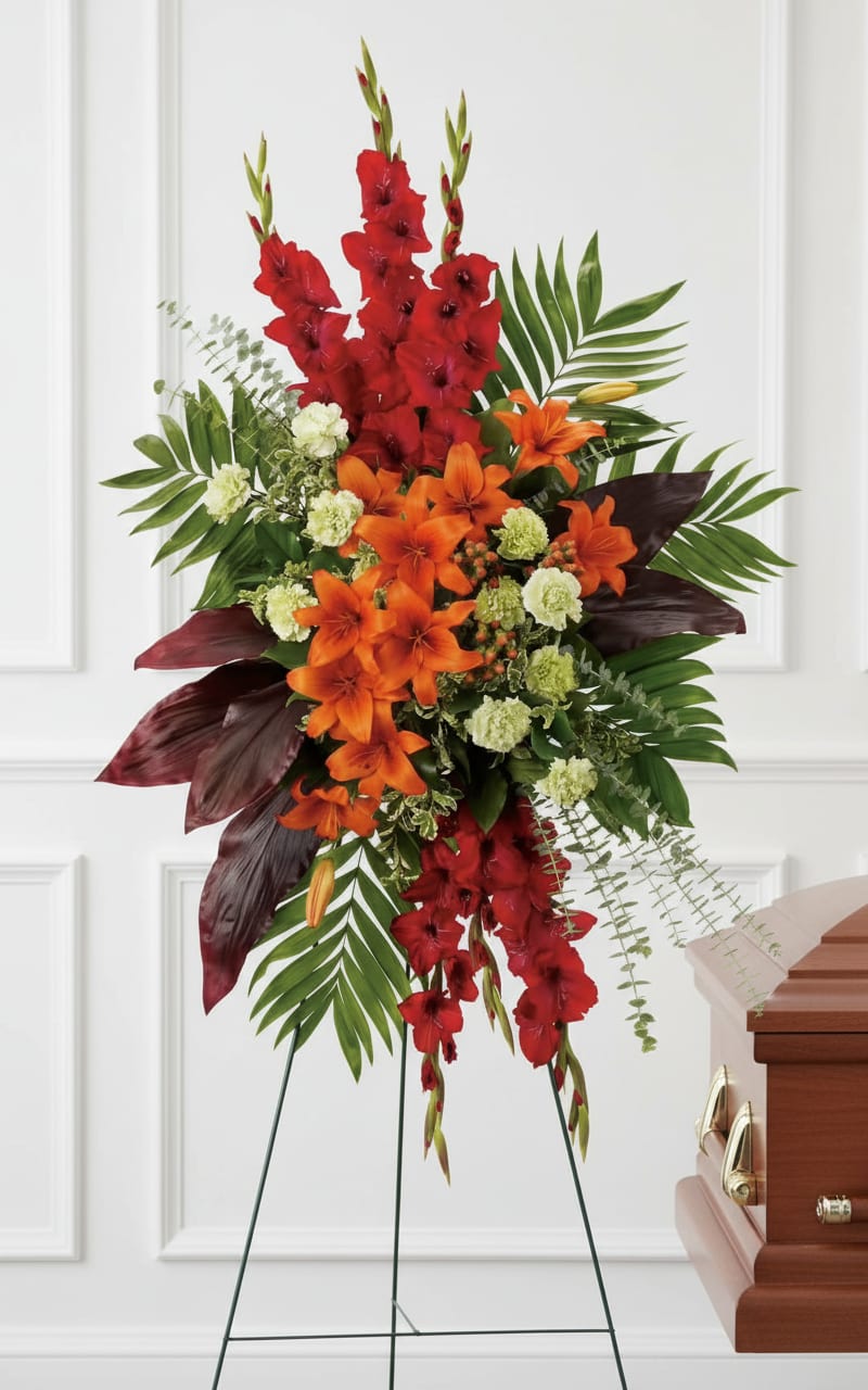 Tall standing spray of red gladiolus, orange lilies, and green carnations on an easel beside a casket.