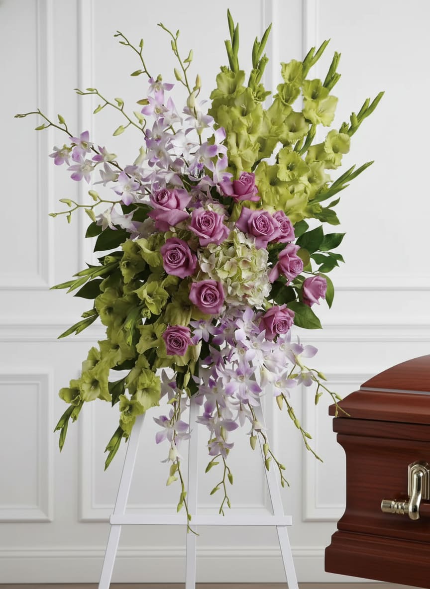 Standing spray of green gladiolus, lavender roses, and white orchids on a white easel beside a casket.
