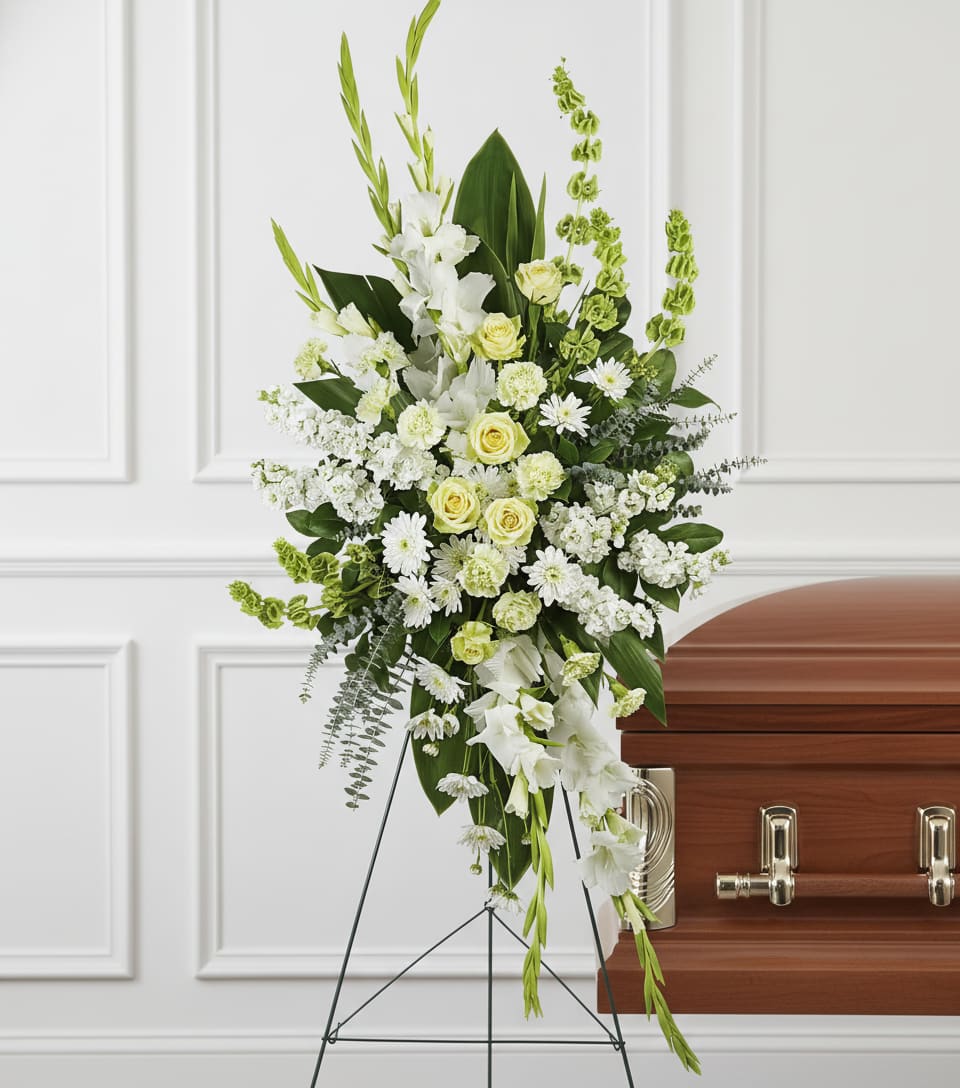 Tall white and pale yellow standing floral spray on an easel beside a wood casket