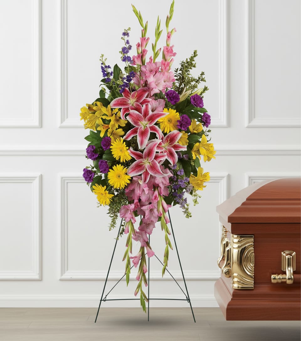 Tall standing spray of pink lilies, yellow daisies, and purple flowers on an easel beside a wooden casket