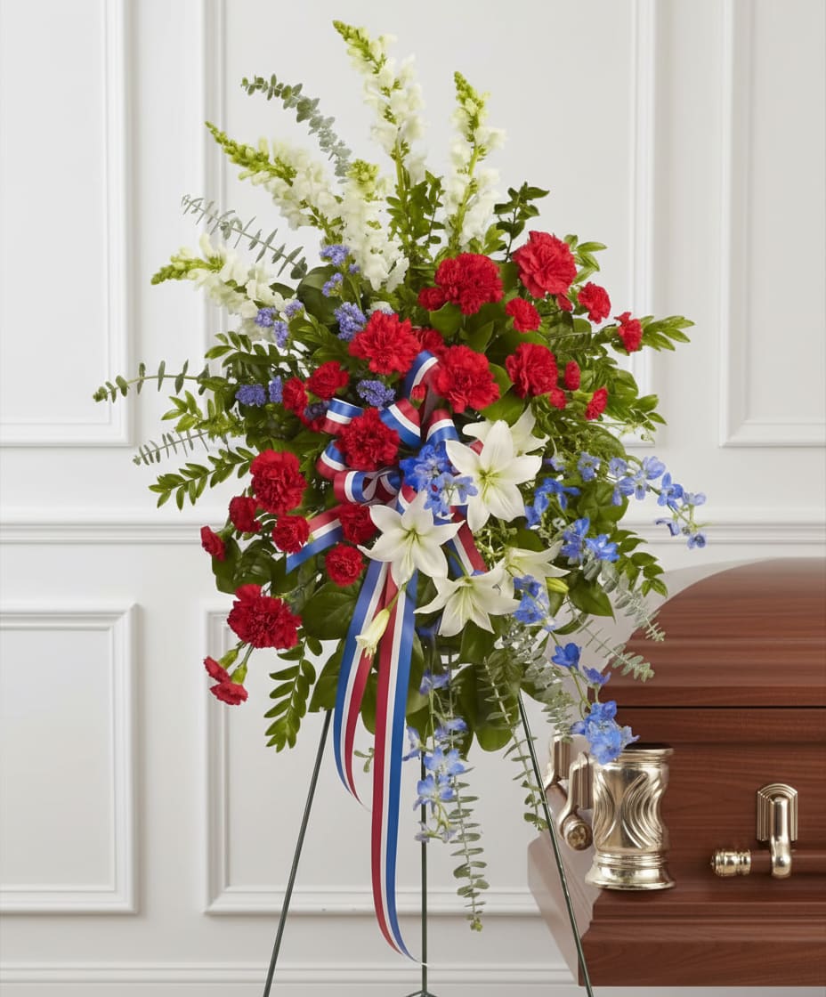 Tall red, white, and blue standing spray with carnations, lilies, and ribbon on an easel beside a casket.