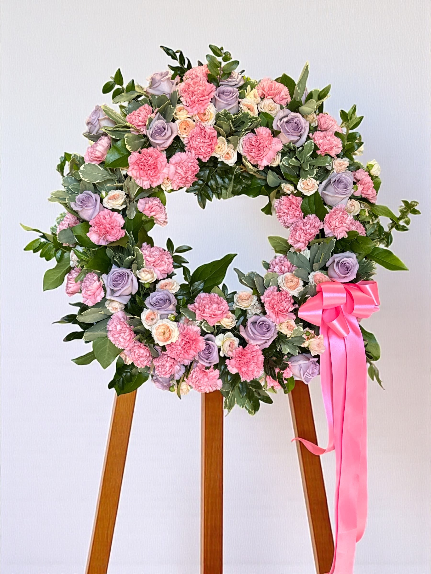 Pink Floral Wreath 18" - Lush greenery and a harmonious blend of soft-colored flowers, including light pink carnations, lavender roses, and small cream spray roses. A large, glossy pink ribbon cascades gracefully from the lower right side of the arrangement, adding a delicate finishing touch.