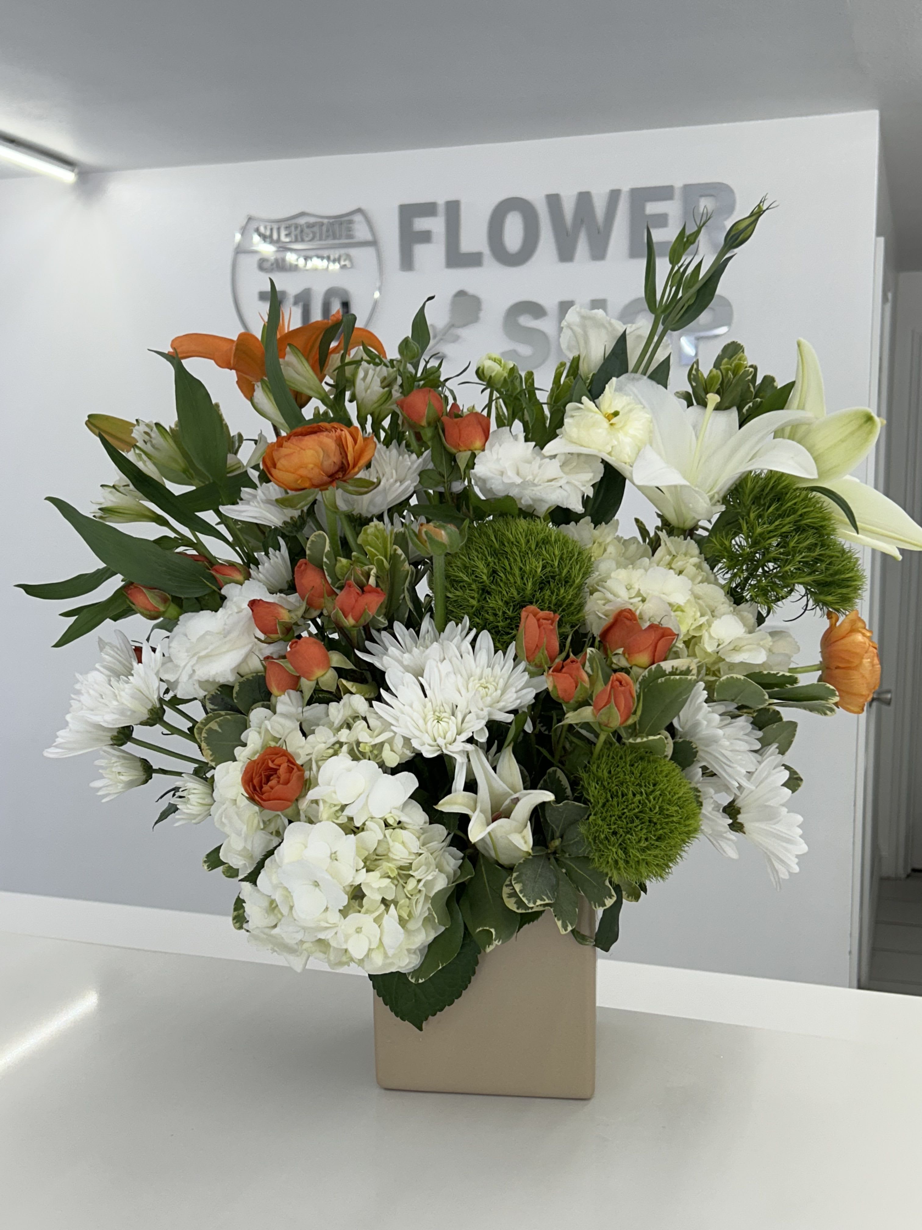 Orange Zest Garden - Bright, fresh, and full of life, Orange Zest Garden is a vibrant arrangement that brings a burst of sunshine into any space. Crisp whites and lush greens create a clean, modern base, while lively pops of orange add warmth and energy. Designed in a sleek container, this piece blends natural texture with a refined, contemporary feel—perfect for celebrations, thoughtful gifts, or simply elevating everyday moments. (If some flowers and/or the vase are not available there will be minor substitutions.)