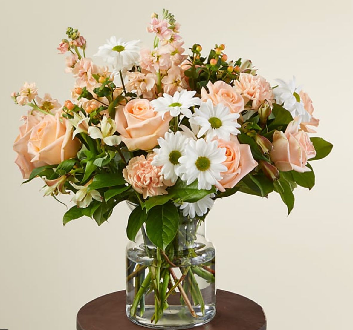 Peach roses, white daisies, and pastel blooms arranged in a clear glass vase.