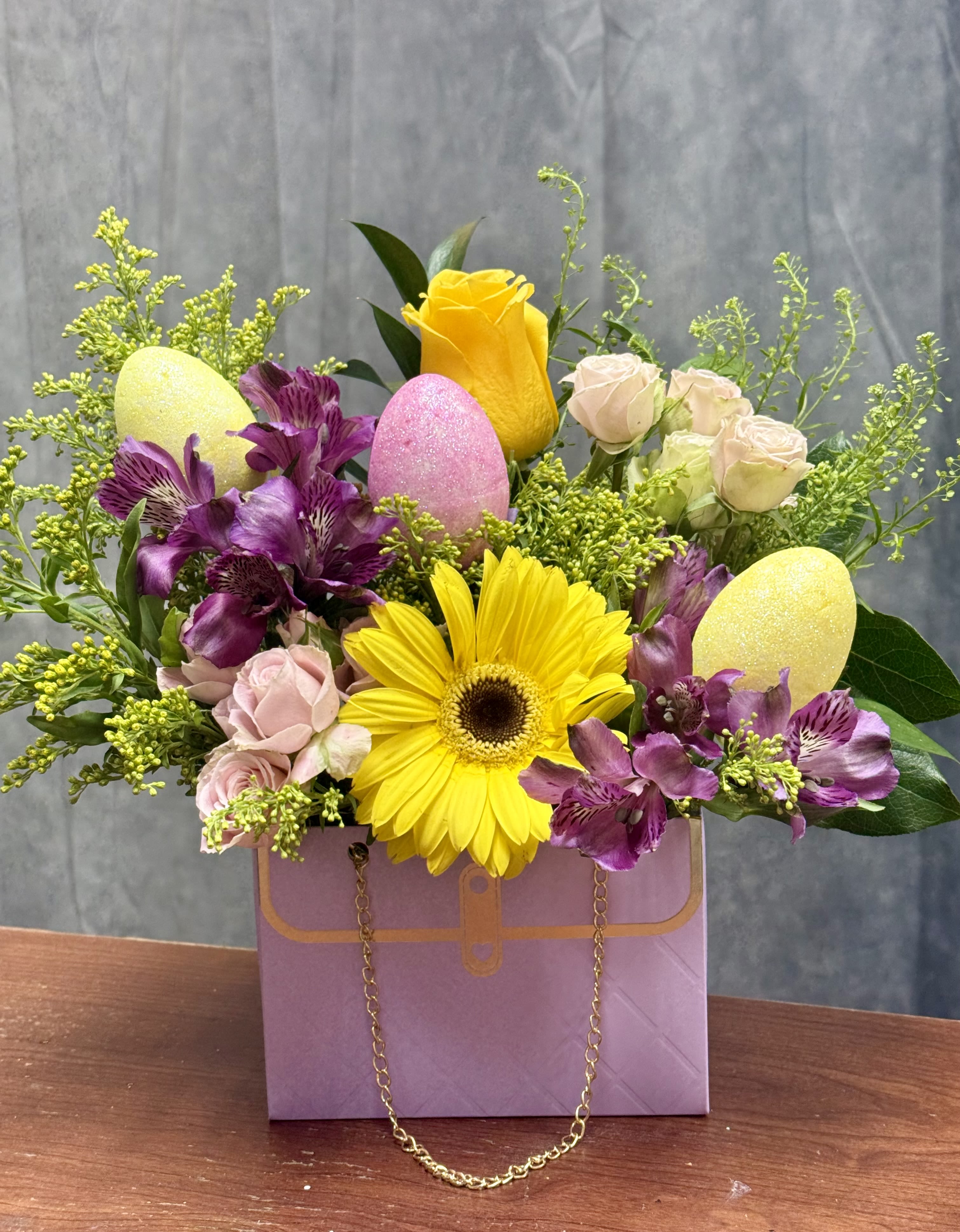 Velvet Garden Purse  - A charming spring design bursting with joy and texture, Velvet Garden features a radiant sunflower, soft blush roses, and vibrant purple blooms, accented with fresh greenery and festive Easter eggs. Arranged in an elegant lavender purse-style box with a delicate gold chain, this piece is the perfect blend of playful and chic—bringing a touch of springtime magic to any space. 