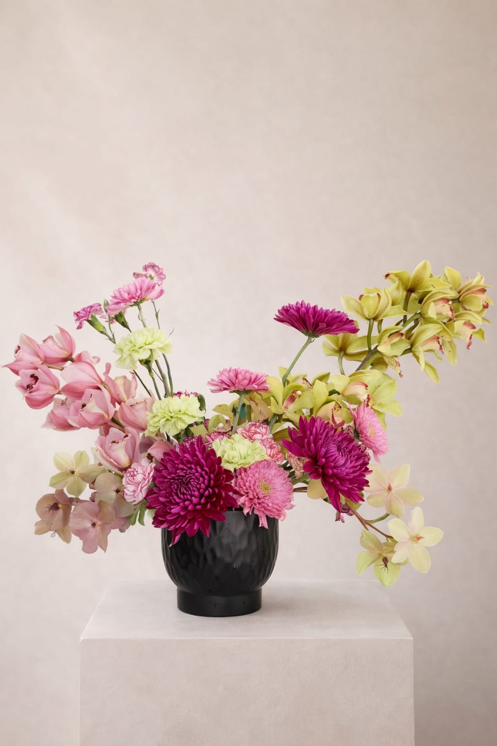 The Cymbidium Symphony  - A fluid, movement-driven arrangement where cymbidium orchids lead the composition with elegance and poise. Cymbidium Symphony showcases arching stems of green orchids that weave through layers of rich magenta and soft pink blooms, creating a harmonious balance of color and form.  Each element is thoughtfully placed to evoke rhythm and flow, with an airy, asymmetrical silhouette that feels both modern and organic. Subtle tonal shifts and textural contrasts add depth, while the overall composition captures a sense of effortless motion.  Refined yet expressive, this piece brings a sophisticated, artful presence to any space.