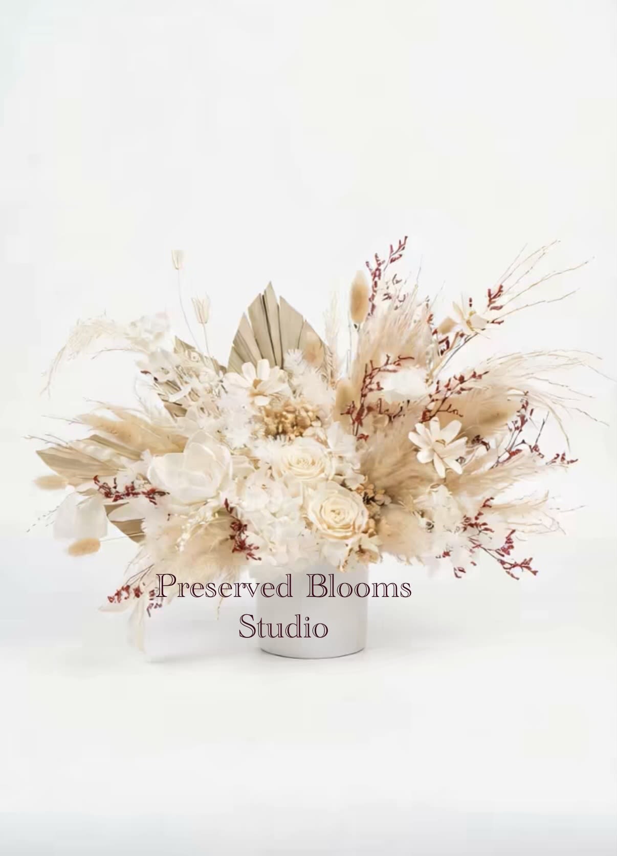 Low arrangement of preserved neutral-toned flowers with roses and dried grasses in a white cylinder vase.