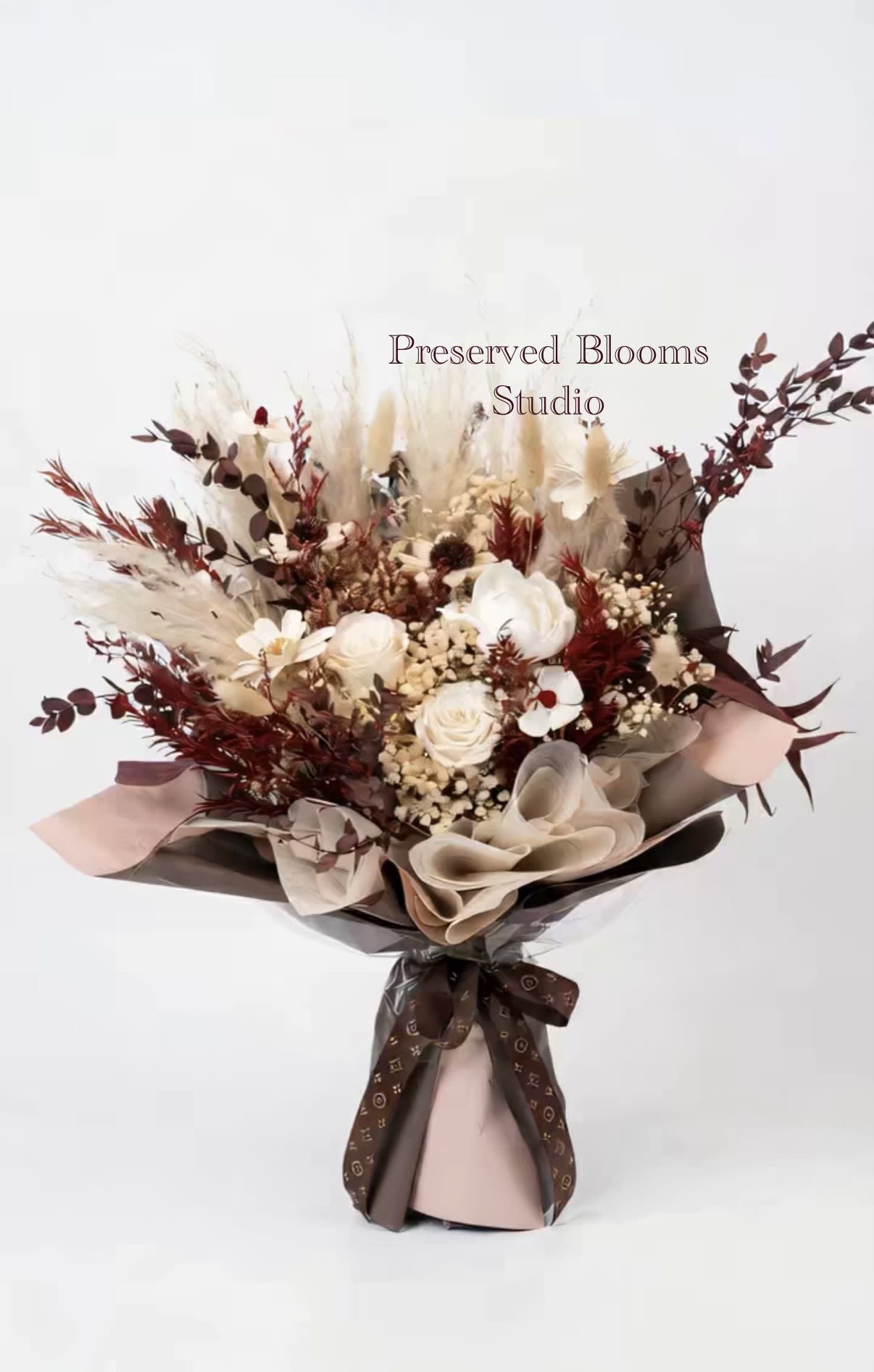 Bouquet of preserved white roses with burgundy dried foliage wrapped in brown and blush paper with a printed ribbon