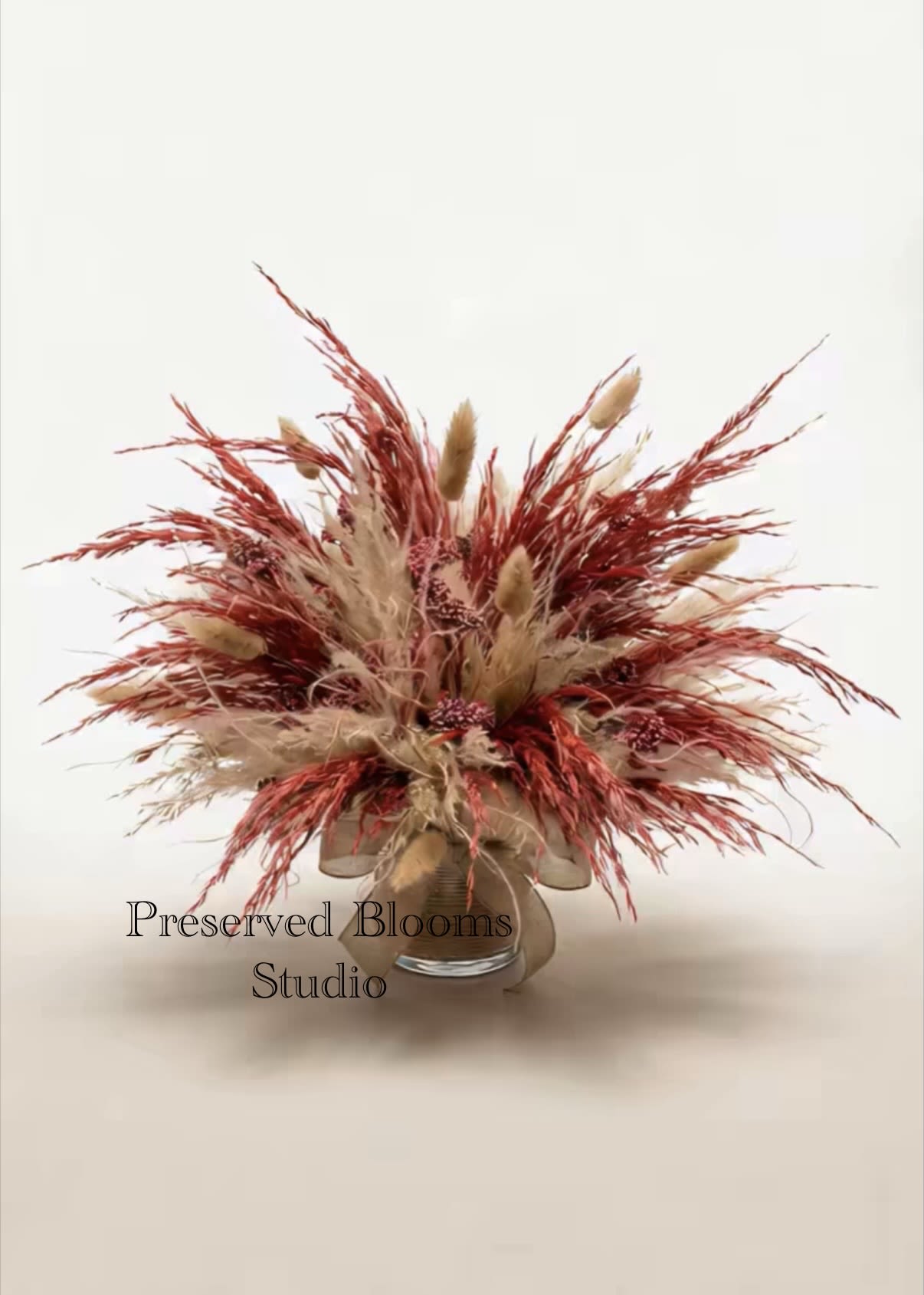 Preserved arrangement of tan and rust dried grasses and seed clusters in a clear glass vase.