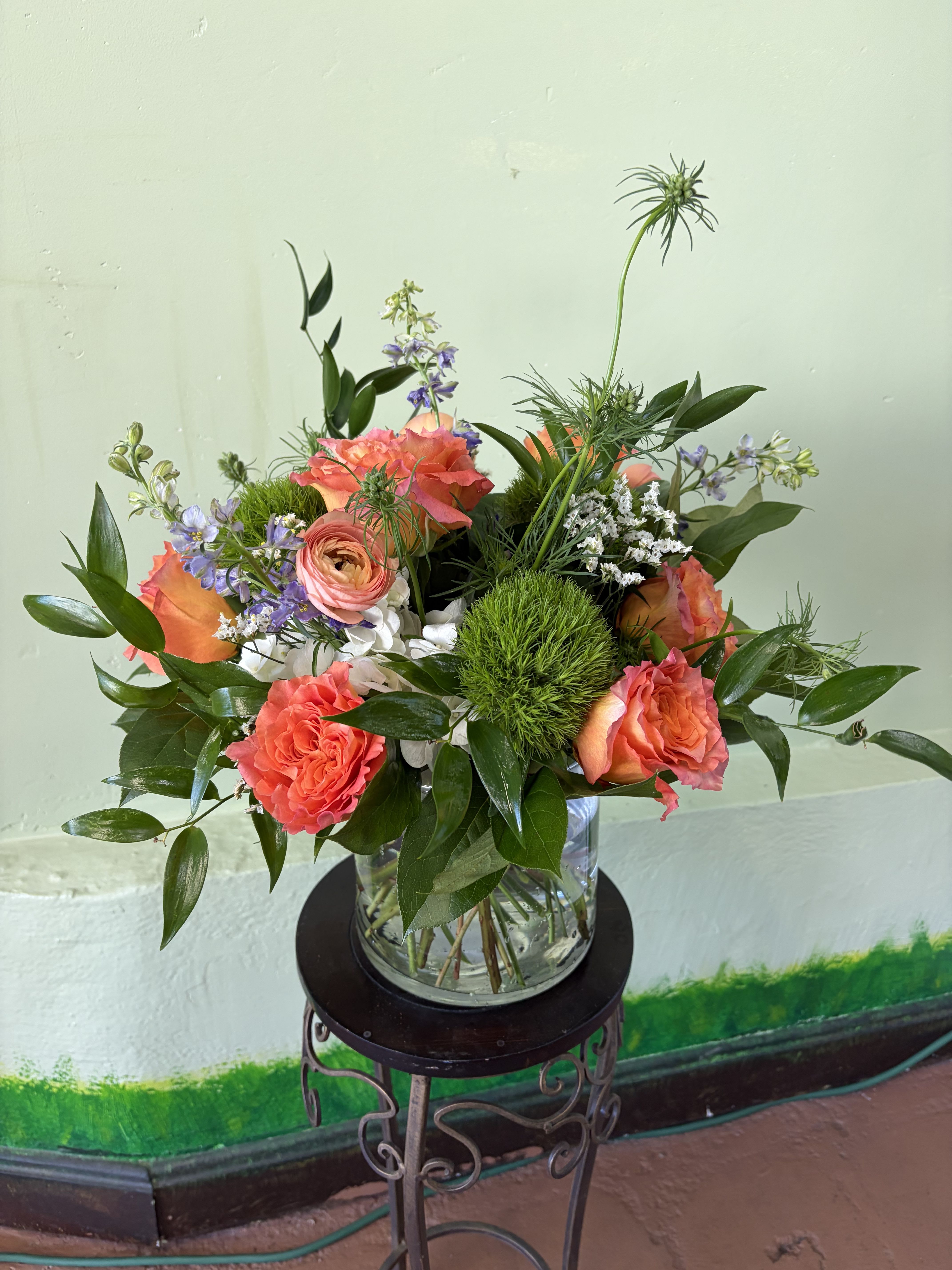 Coral roses, ranunculus, and green dianthus in a clear glass vase on a metal stand