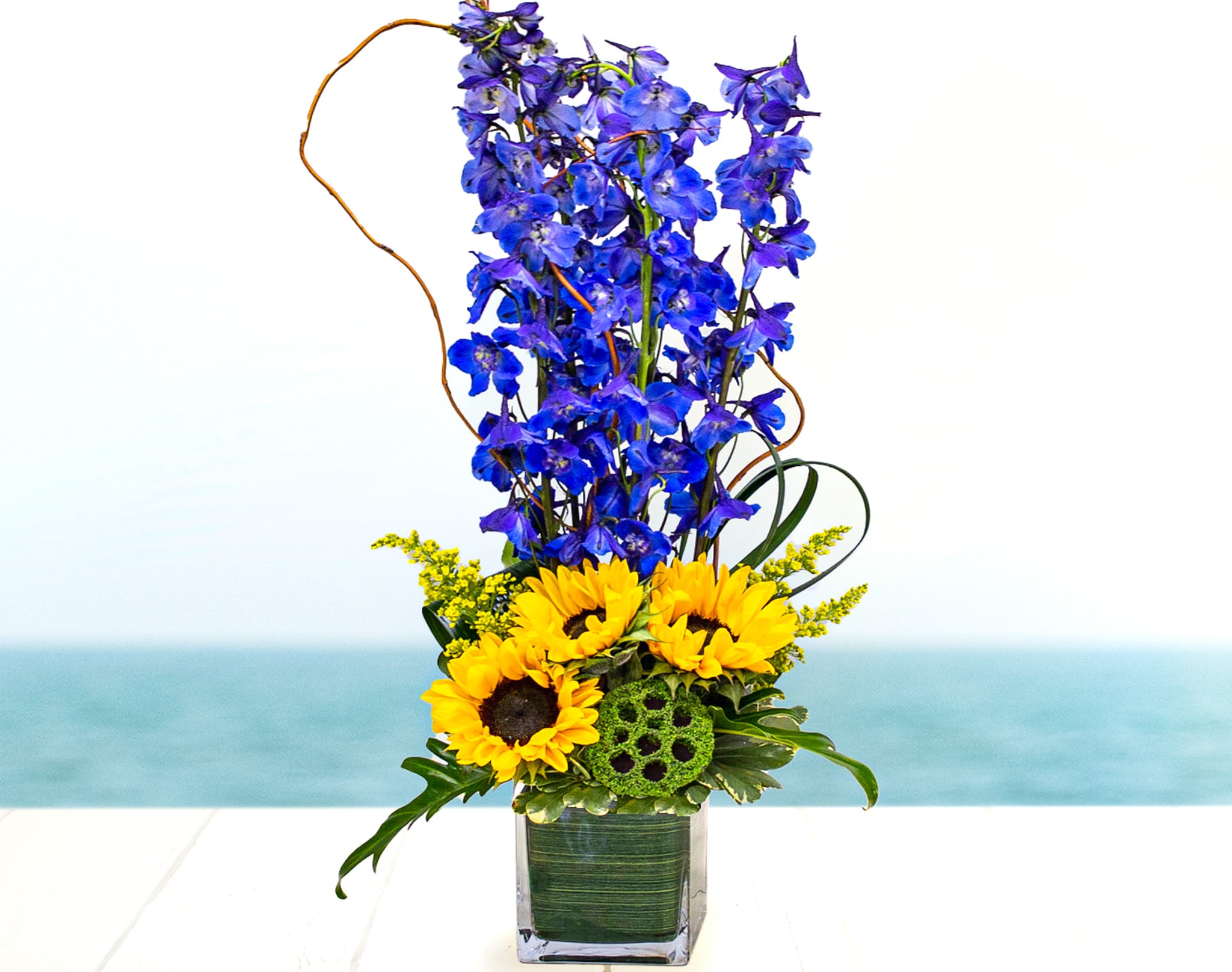 Tall blue delphinium and yellow sunflowers arranged in a clear rectangular glass vase