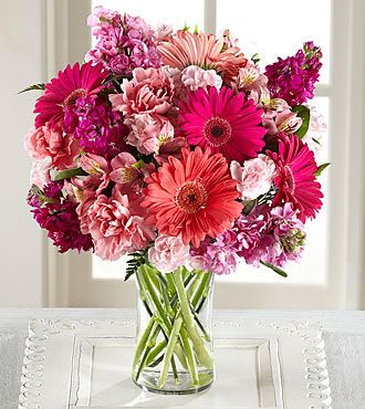 Blushing Beauty  - A lush arrangement bursting with vibrant pinks and soft blush tones, this design features a beautiful mix of gerbera daisies, carnations, and seasonal accent blooms. Arranged in a clear glass vase, the fresh greenery and layered textures create a full, eye-catching look perfect for celebrations, gifts, or brightening any space.  Flowers may vary slightly based on availability, but the overall color palette and style will be thoughtfully preserved.
