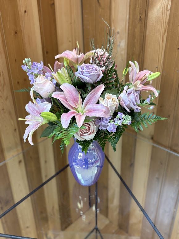 Lavender Whisper - Lavender Whisper opens with a breath of pastel romance-pale pink lilies and lavender-hued roses mingle with soft blush roses to create a birthday bouquet, anniversary arrangement, or Mother's Day gift that feels like a gentle embrace. Perfect for Easter or spring celebrations yet lovely year-round, this floral arrangement speaks to someone you want to comfort, celebrate, or simply make feel cherished: Mother, friend, new parent, or the person recovering at home.  This design pairs light purple stock and airy fillers with lush greenery for texture and movement, all gathered in a glossy purple glass vase that echoes the lavender palette and lends a modern, gift-ready finish. The lilies bring bold, open petals and soft scent while the roses and stock add layered color-pink, lavender, purple, and white-for an airy, feminine look ideal for housewarming, get well, thank you, thinking of you, I'm sorry, or just because moments.  Order with confidence from Haehn Florist, Greenhouses, &amp; Flower Delivery at 410 Hamilton Rd. Wapakoneta; substitutions may be made for seasonal availability but will preserve the intended color story and style. Visit our Facebook page at www.facebook.com/haehnflorist for updates and seasonal specials.