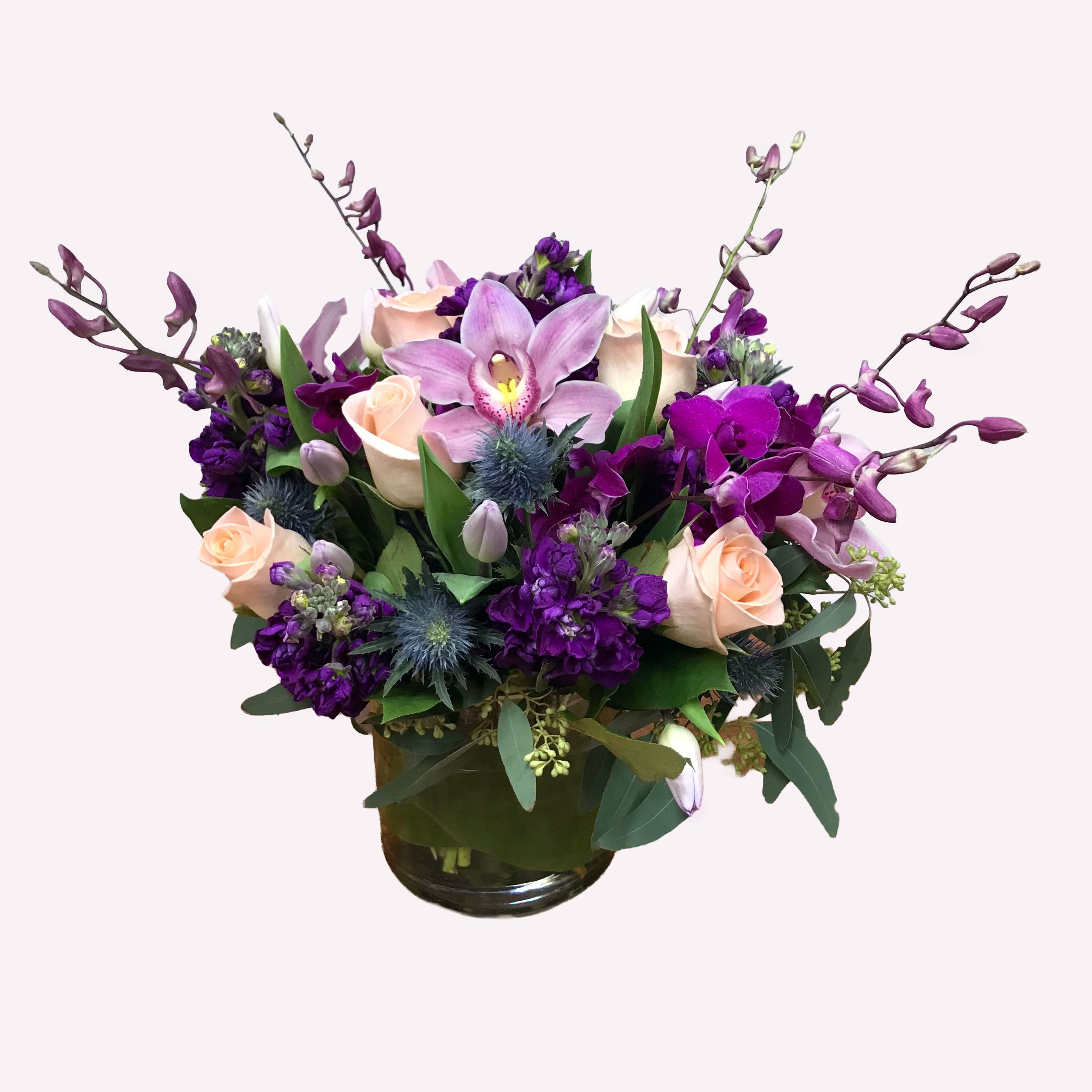 Crown's Jewel - Are you celebrating a particular royal in your life? This arrangement is the perfect match for you. An array of vibrant purples dendrobium orchids and stocks, soft lavenders cymbidium orchids and tulips (seasonal), pale peach roses, thistle, and seeded eucalyptus completes the look. Flowers arranged in a short cylinder vase with tropical green leaf.
