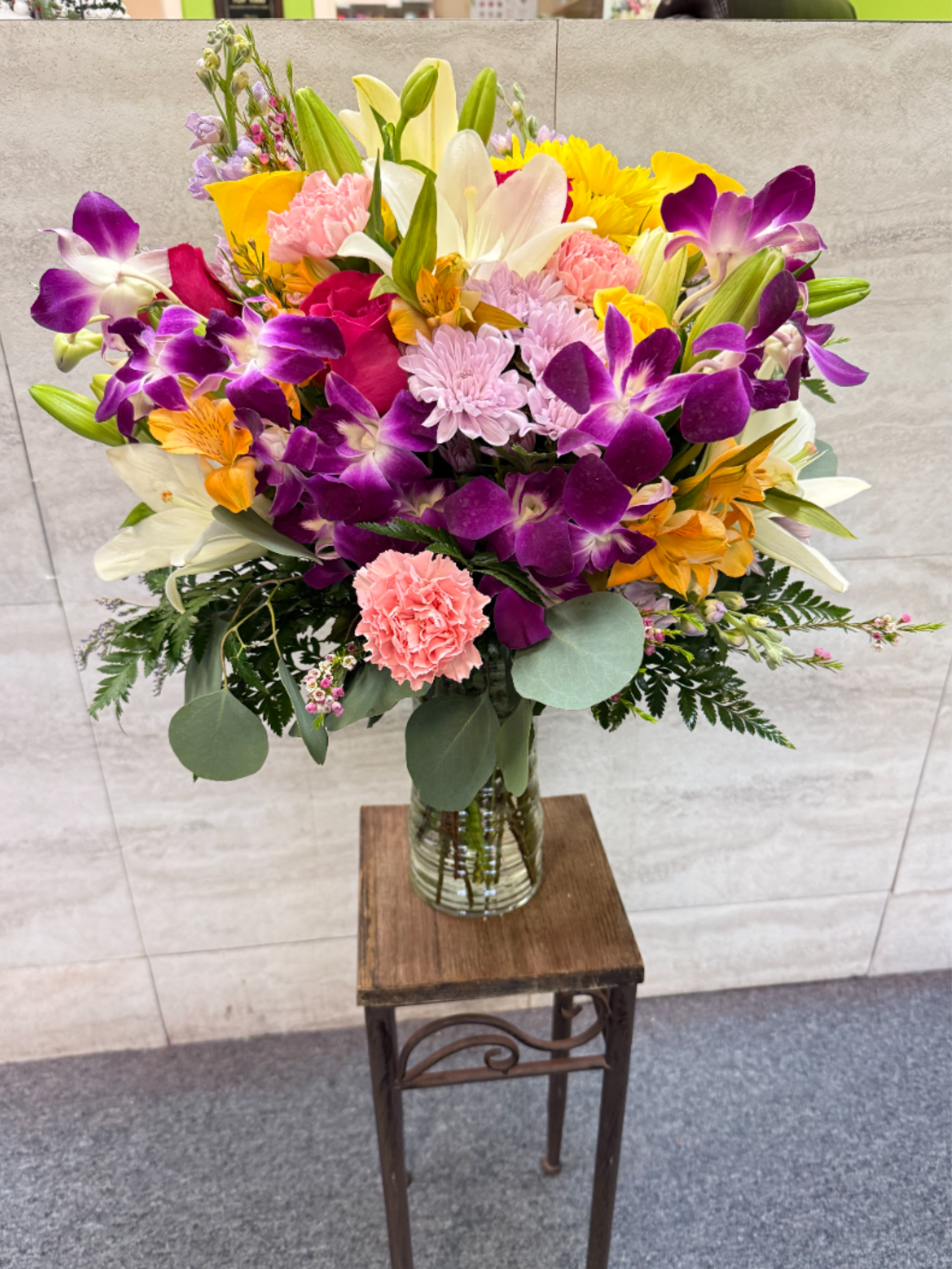 Lani Bloom - A vibrant, tropical-inspired bouquet featuring stunning dendros, elegant lilies, and soft carnations, accented with fresh greenery. Its bright mix of blooms creates a lively, island-inspired feel—perfect for bringing a touch of paradise to any space. 