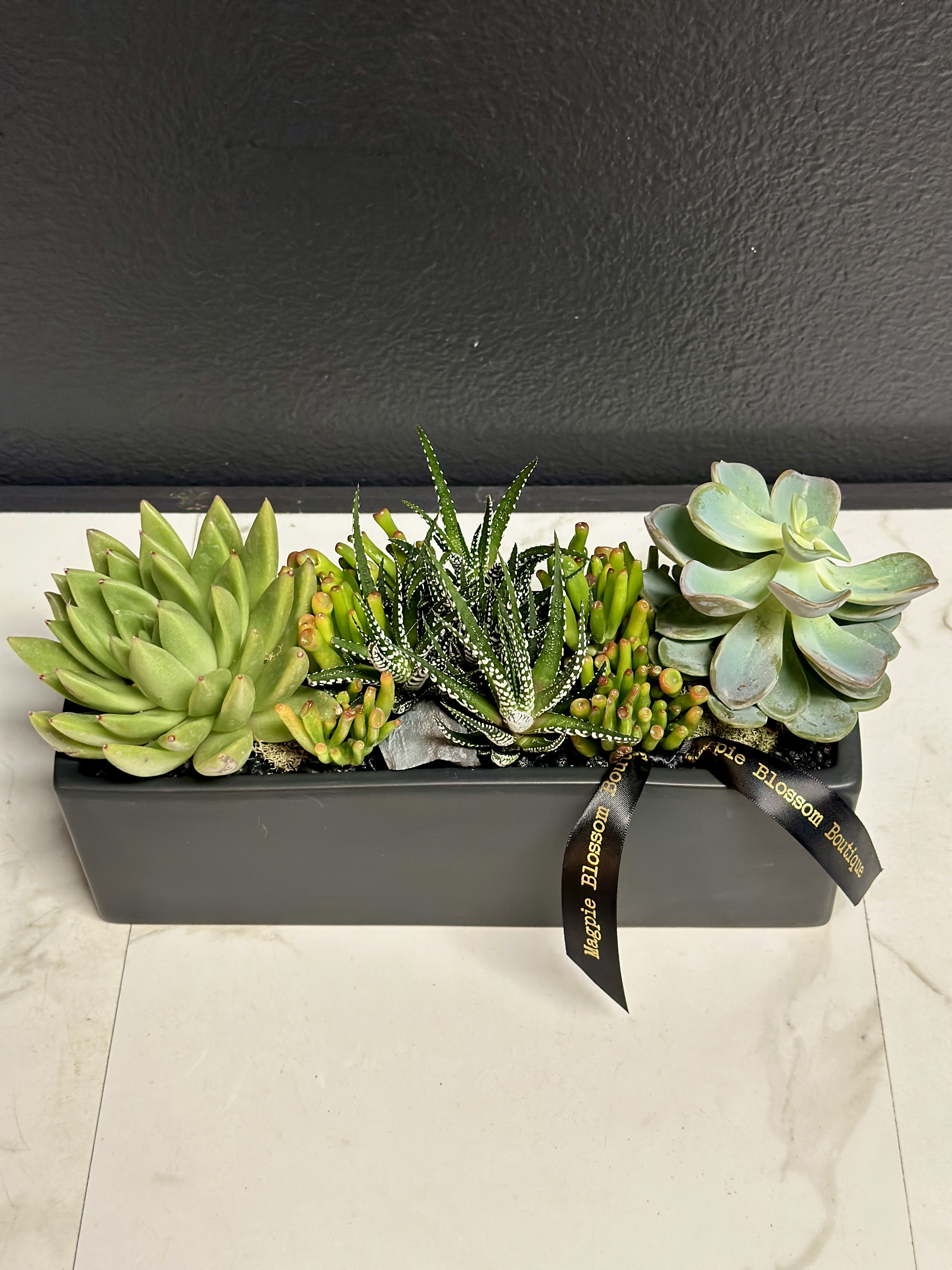 The Nero - Add a touch of contemporary charm to any space with our Nero Garden. A stunning arrangement of succulents is enhanced with black pebbles, and stone accents.  Dimensions | Approximately 12x7"