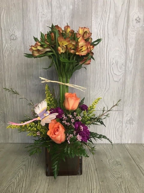 Tall mixed arrangement with orange roses, purple flowers, and alstroemeria in a brown glass cube vase
