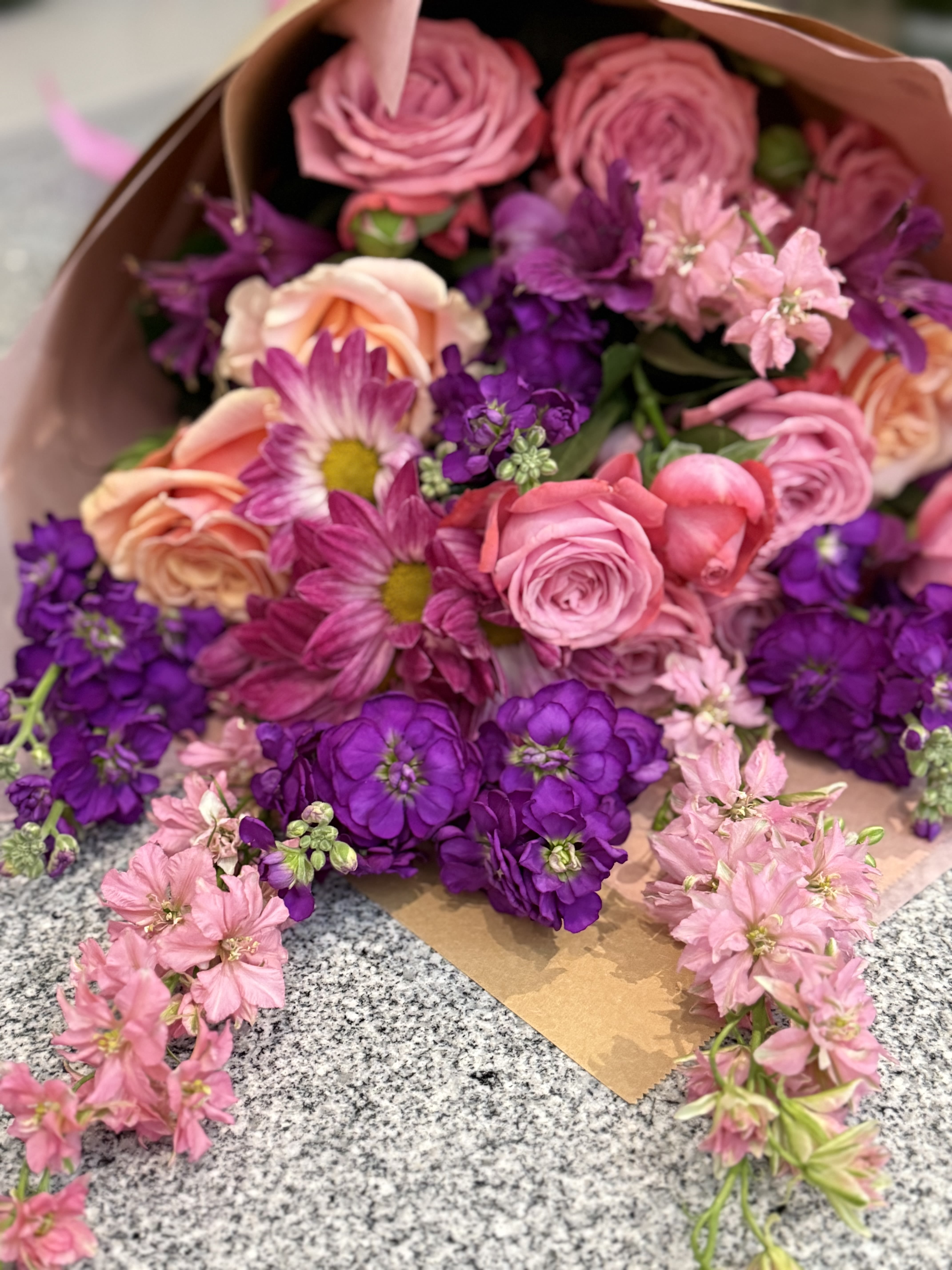Wrapped bouquet of pink and peach roses with purple and pink blooms on a granite surface
