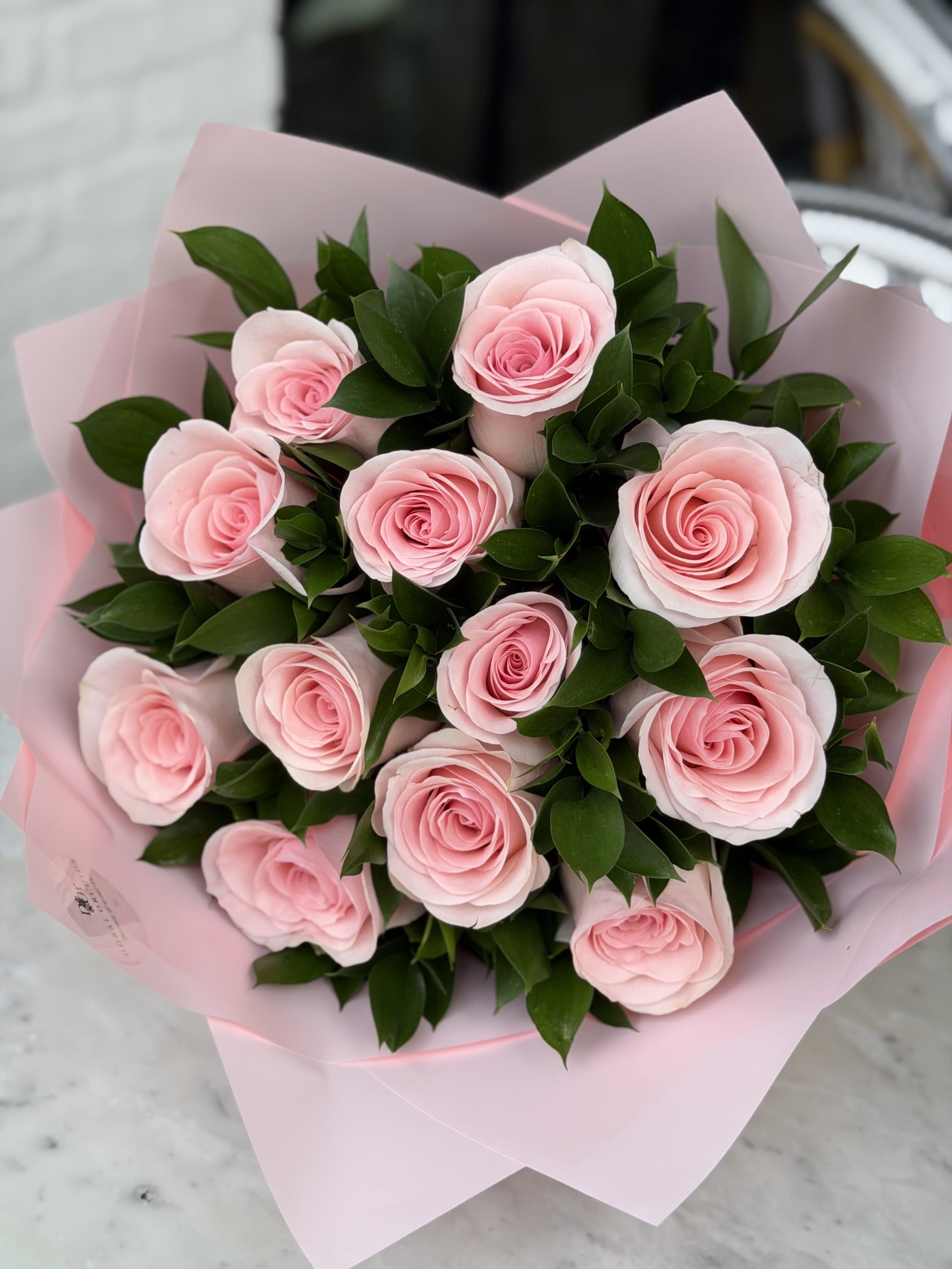 Pink Dozen - A romantic and elegant arrangement featuring a dozen soft pink roses paired with delicate ruscus greenery, wrapped in luxurious blush pink paper. The fresh roses are hand-selected for their premium quality and beautifully arranged to create a full, balanced bouquet with a soft, feminine feel. Perfect for celebrating love, appreciation, or any special moment, this bouquet offers a timeless and graceful presentation with a modern, refined touch.