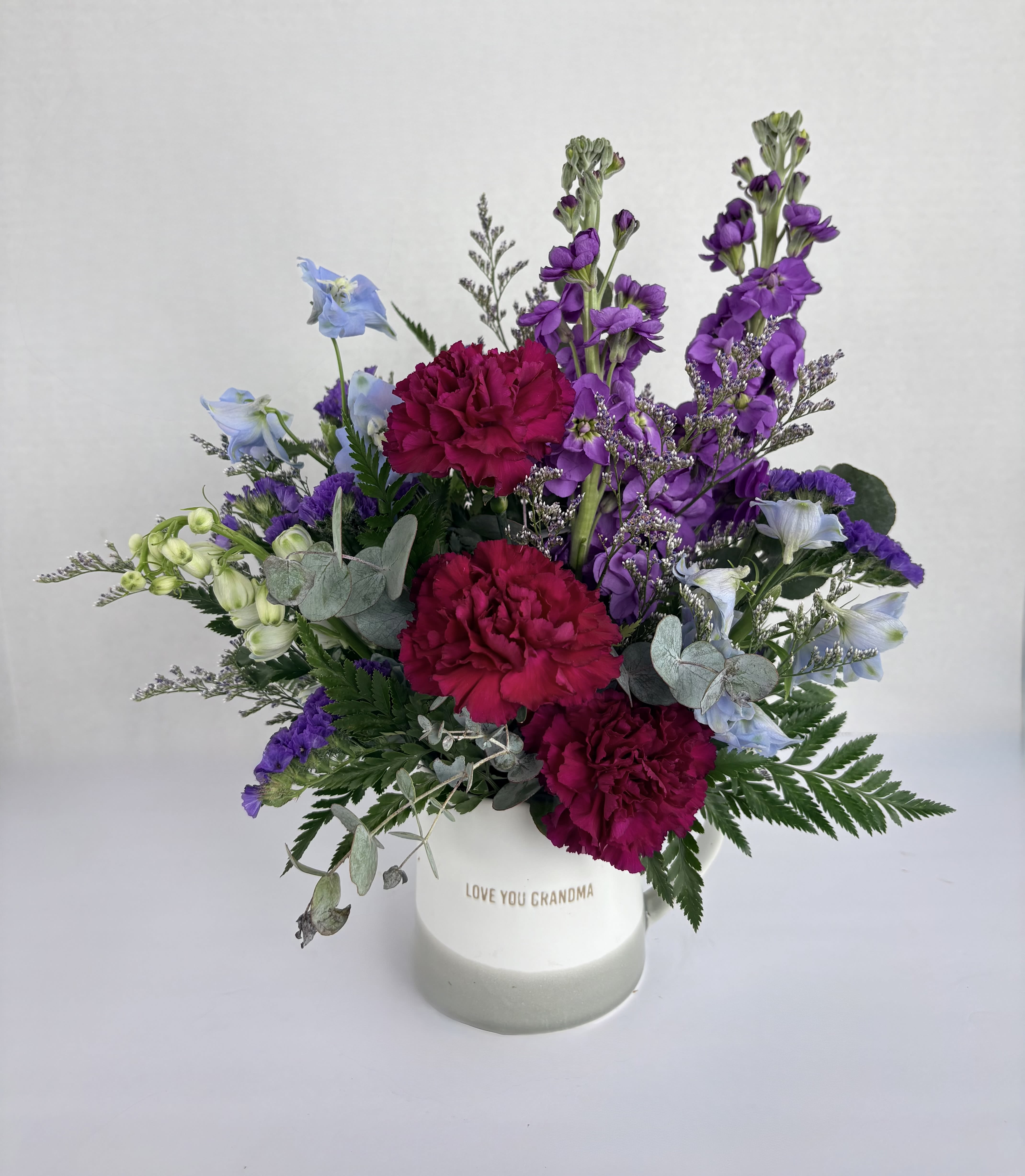 Grandma's Garden  - Grandma's Garden is designed in a mug with "Love you Grandma" etched into the front. This simple mug is enhanced with magenta carnations, purple stock, blue delphinium, purple statice, and limonium. Give the gift of love through flowers with Grandma's Garden. 