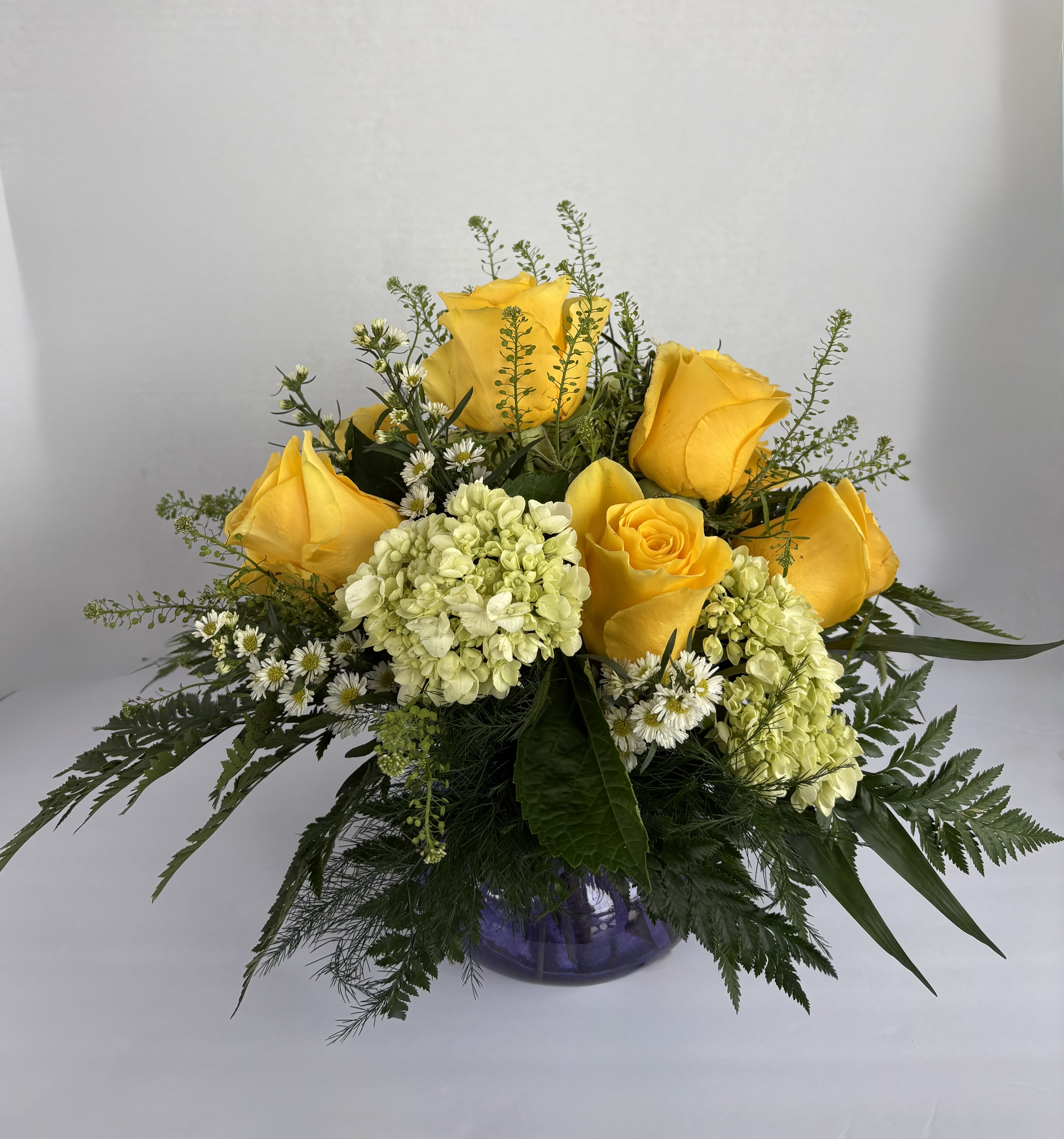 Regal Roses  - This noble arrangement includes seven yellow roses with three green hydrangeas, designed in a small colored vase. Adorned with penny cress and white monte casino, this bright design will add a sprinkle of regality to any event or occasion. 