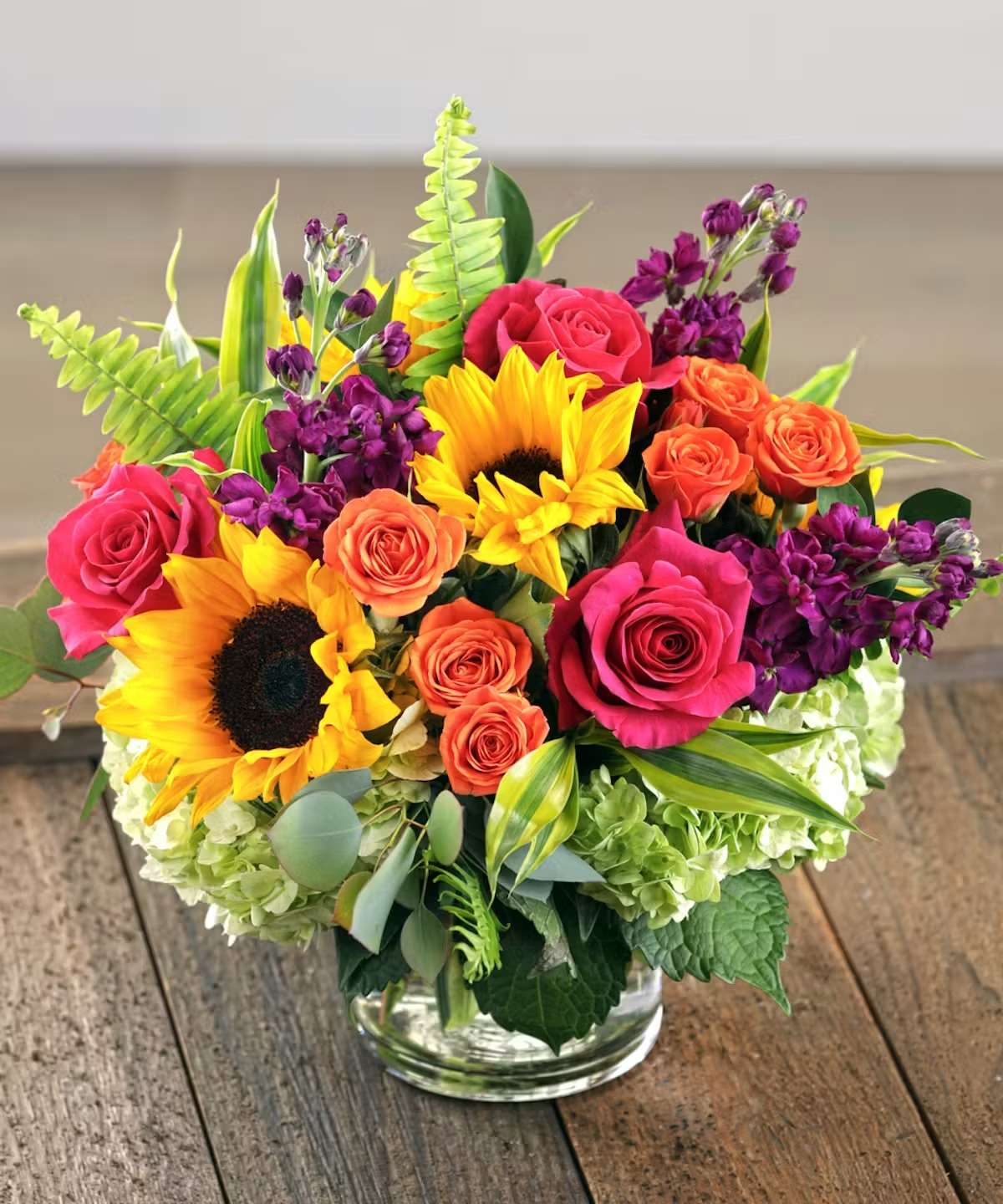 Vibrant Expression - Brighten any room with this cheerful arrangement featuring vibrant sunflowers, hot pink roses, orange spray roses, purple stock, and fresh seasonal greenery in a clear glass vase. Bursting with bold color and garden-inspired charm, it’s the perfect gift to celebrate a birthday, send encouragement, or simply make someone smile.  Flowers may vary slightly based on availability, but the overall color palette and style will be thoughtfully preserved.