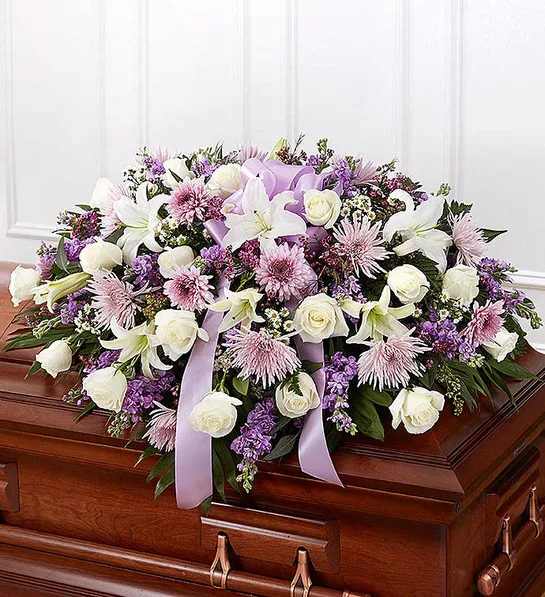 Lavender &amp; White Mixed Half Casket Cover - Half casket cover arrangement of white roses, hybrid lilies and monte casino, lavender cremones, spider mums and stock, purple waxflower and fresh greenery; accented with a lavender satin ribbon Arrangement measures approximately 16"H x 38"L x 26"D Traditionally sent by the immediate family to the funeral home Our florists use only the freshest flowers available, so colors and varieties may vary