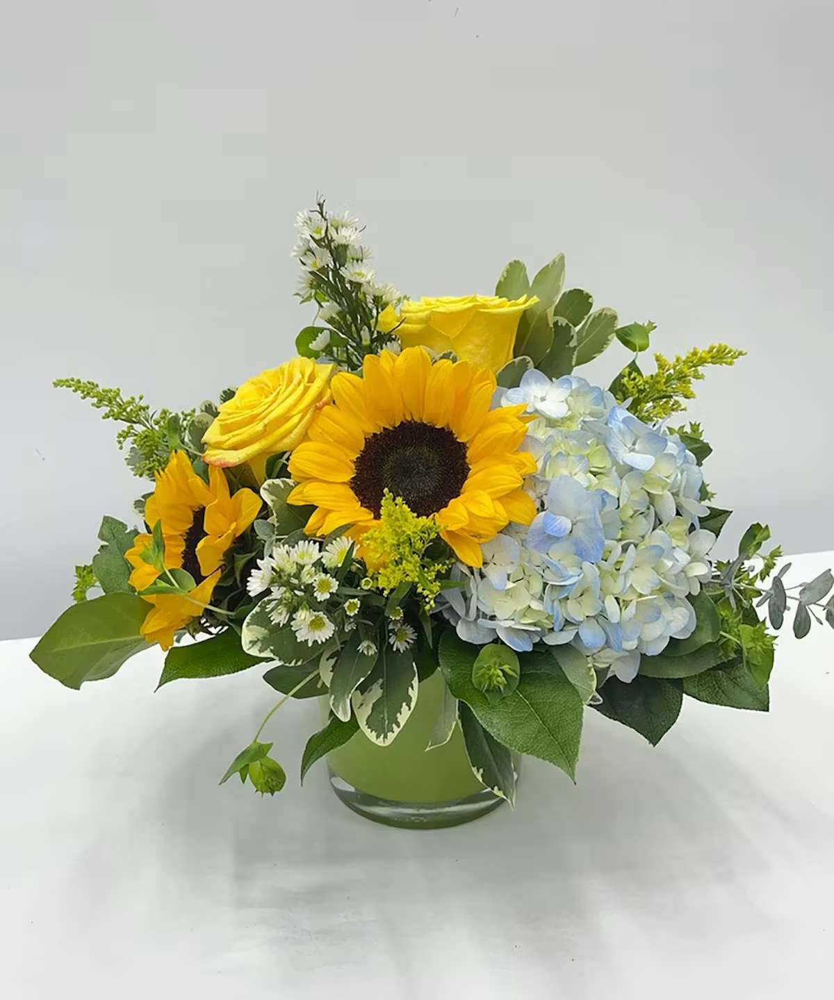 Sunlit Serenity - A fresh and cheerful arrangement featuring radiant sunflowers, sunny yellow roses, soft blue hydrangea, delicate white accent flowers, and lush greenery gathered in a clear glass vase. With its bright, uplifting colors and garden-inspired style, Sunlit Serenity is a beautiful choice for birthdays, get well wishes, thank you gifts, or simply to brighten someone’s day.  Disclaimer: Flower varieties and colors may vary based on seasonal availability. Substitutions may be made with flowers of equal or greater value while maintaining the overall look and feel of the arrangement.