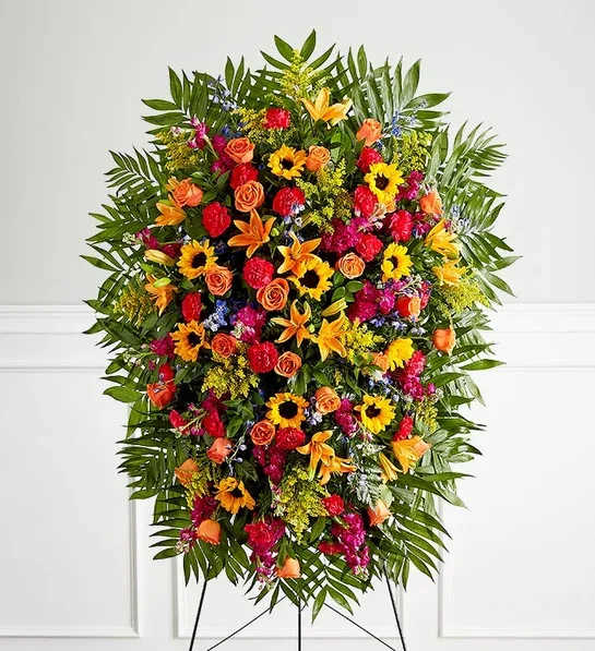 Vibrant Funeral Standing Spray - One-sided standing spray arrangement with orange roses and Asiatic lilies, golden sunflowers, blue delphinium, fuchsia stock and hot pink carnations; accented with assorted greenery Large measures approximately 56"H x 38"W without stand Medium measures approximately 46"H x 34"W without stand Arrives on an easel Appropriate for the funeral home or gravesite Our florists hand-design each arrangement, so colors and varieties may vary due to local availability To ensure lasting beauty, Asiatic lilies may arrive in bud form and will fully bloom over the next few days