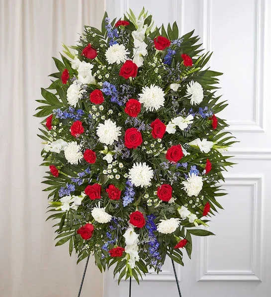 Red, White &amp; Blue Sympathy Standing Spray - Standing spray arrangement with red roses and carnations; white football mums, gladiola and monte casino; blue delphinium; purple limonium; accented with soft, lush greenery and satin ribbon Measures approximately 46"H x 34"L  Measures approximately 36"H x 26"M   without stand Arrives on an easel Appropriate for the funeral home or gravesite Our florists hand-design each arrangement, so colors and varieties may vary due to local availability