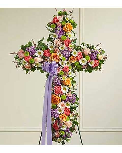 Pastel Mix Cross - A bright and beautiful display of the celebration of life with a stunning mix of light and pastel florals including roses carnations chrysanthemums and mixed greens adorned with a soft purple bow and mounted upright on an easel.