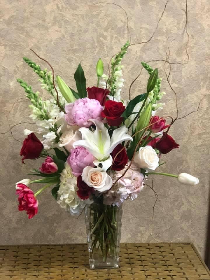 Tall mixed arrangement of pink peonies, red and white roses, lilies and tulips in a clear glass vase
