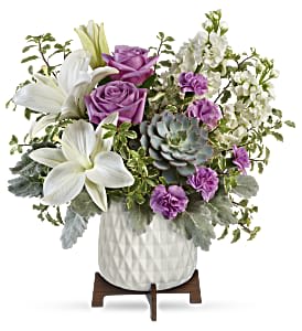 Arrangement of white lilies, lavender roses, purple carnations and a succulent in a white pot on wooden stand