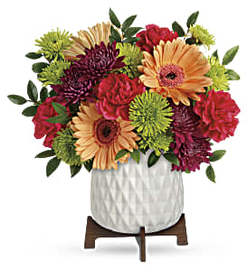 Compact bouquet of orange gerbera daisies, red carnations, and green mums in a white textured pot on a wooden stand