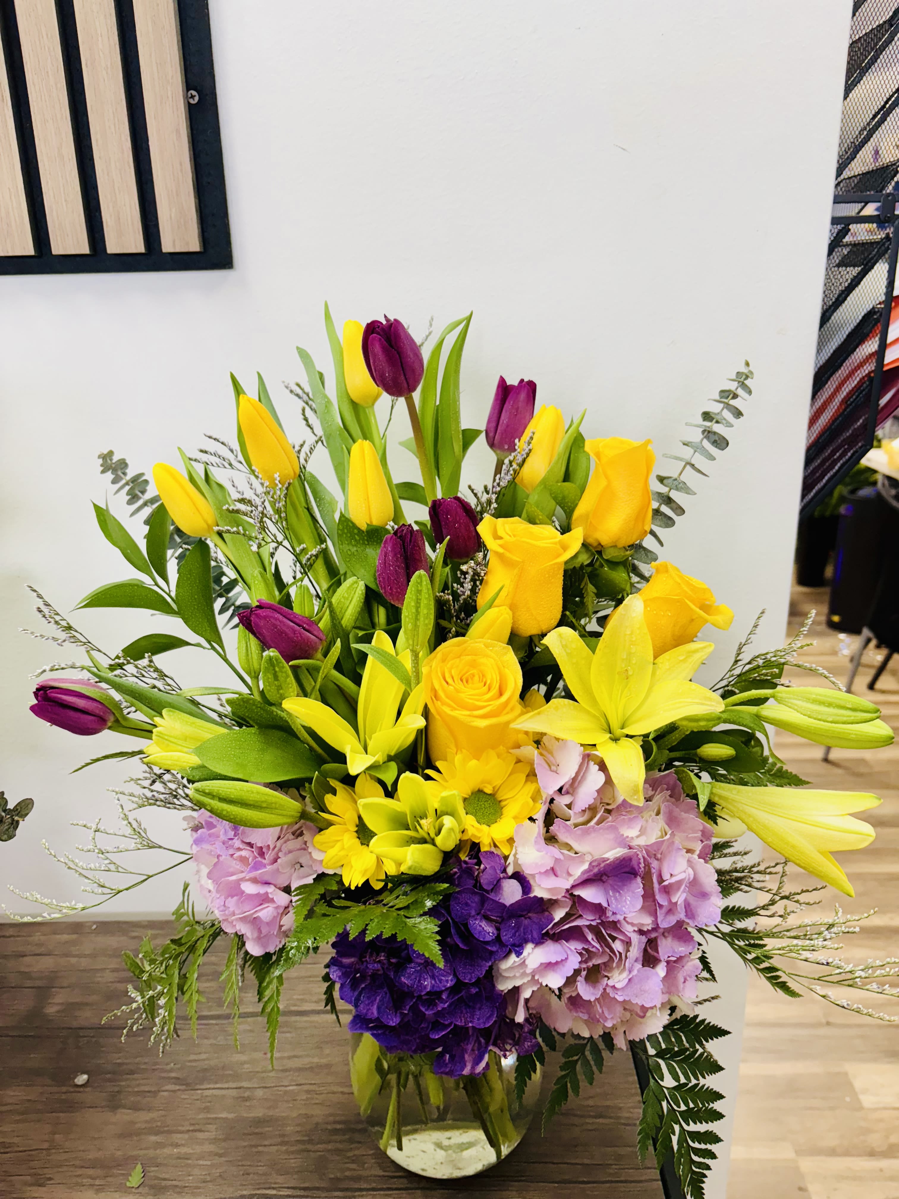 Rayos de Primavera - Vibrant arrangement featuring yellow roses, yellow lilies, daisies, purple tulips, and hydrangeas in purple and lavender tones, arranged in a clear glass vase. A bright, fresh design perfect for birthdays, thank‑yous, or uplifting any space