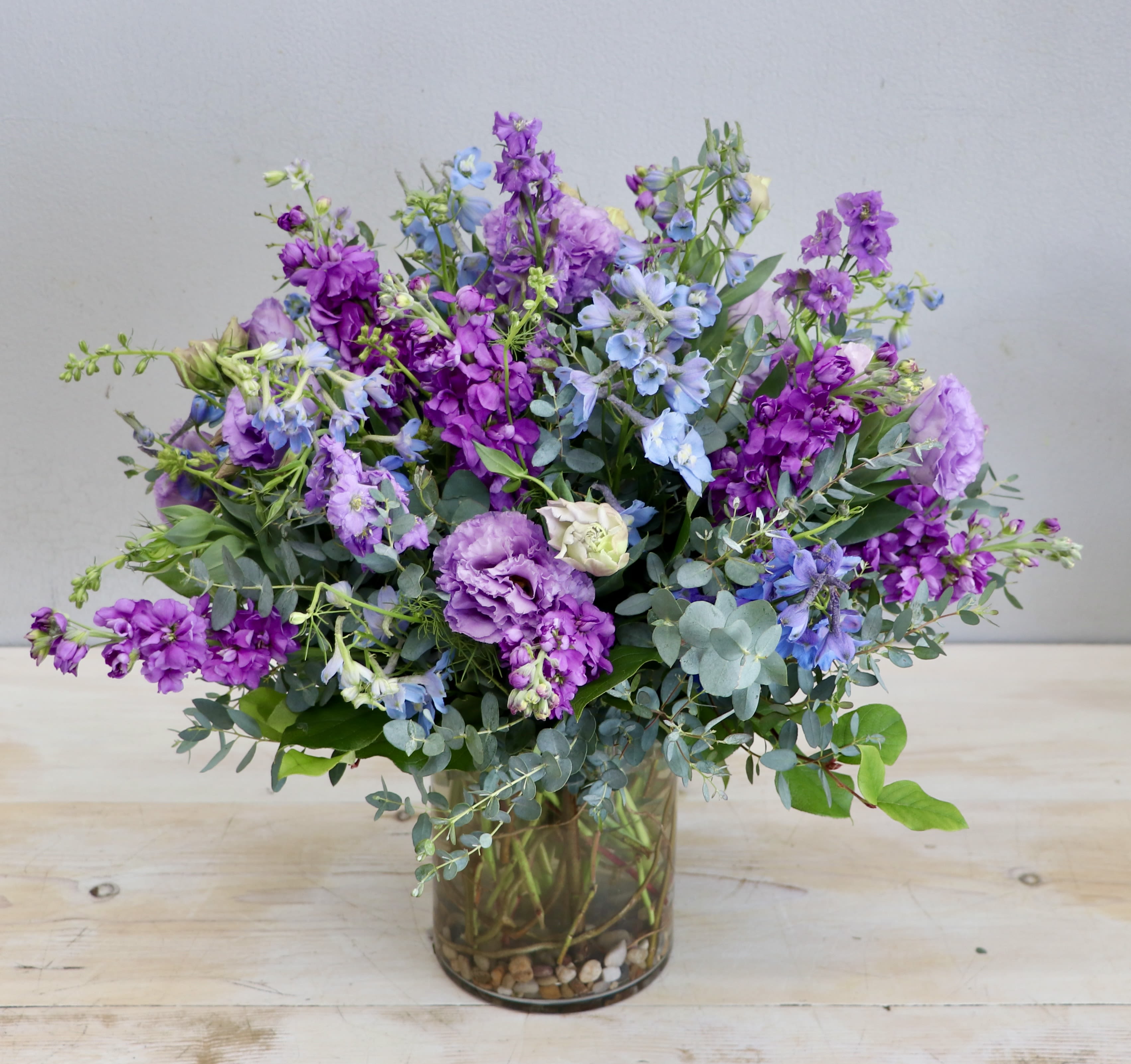 Wild Violet Skies - My Glendale Florist  - This arrangement features baby blue delphiniums with lavender lisianthus and purple stock in a glass container, accompanied with seasonal greenery. Don't forget to upgrade for more blooms! 