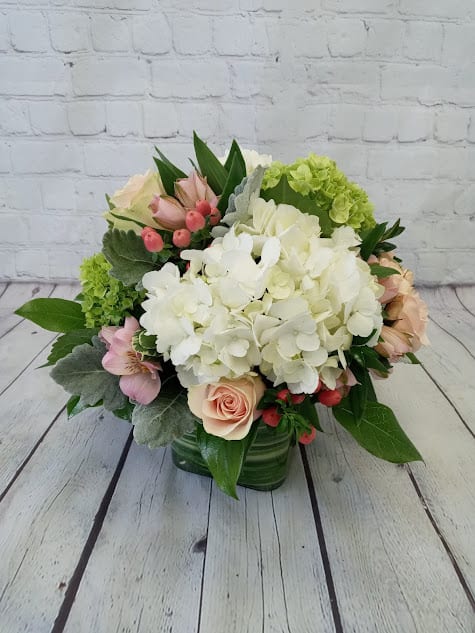 Low arrangement of white hydrangeas with peach roses and pink accents in a green glass cube vase