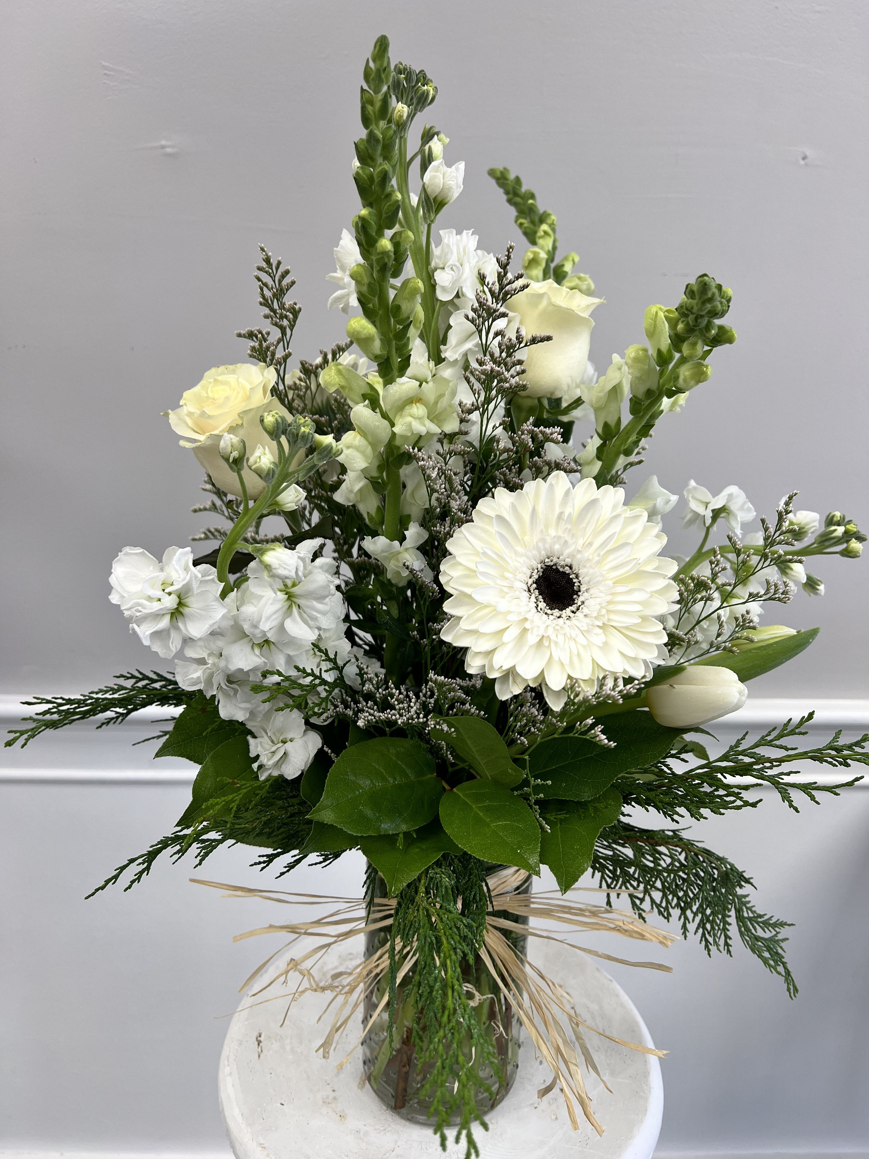Tall white arrangement with roses, gerbera daisy, snapdragons, and tulips in a clear glass vase tied with raffia.