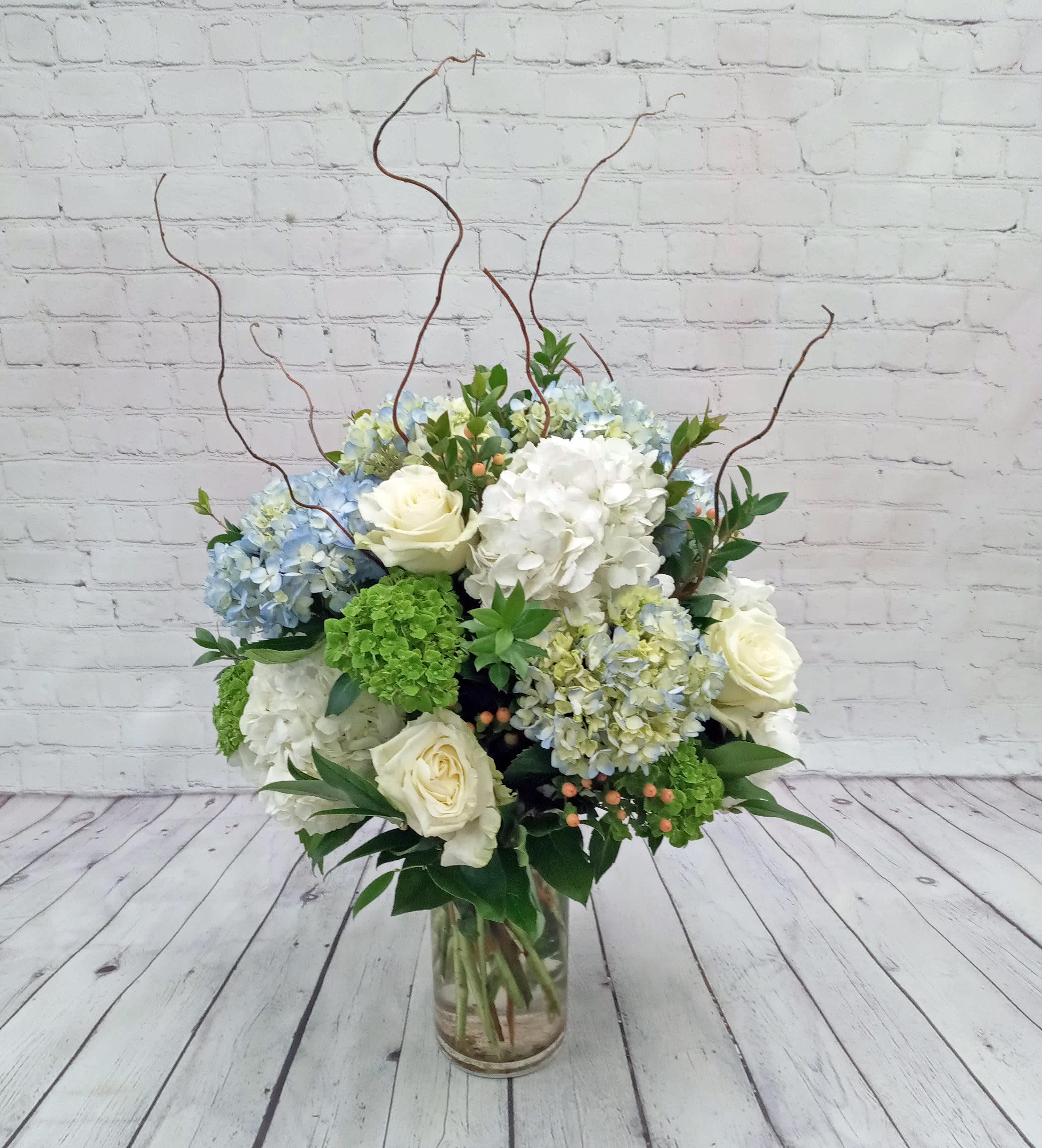 Tall glass vase arrangement with white roses, blue and white hydrangeas, green blooms, and curly willow branches.