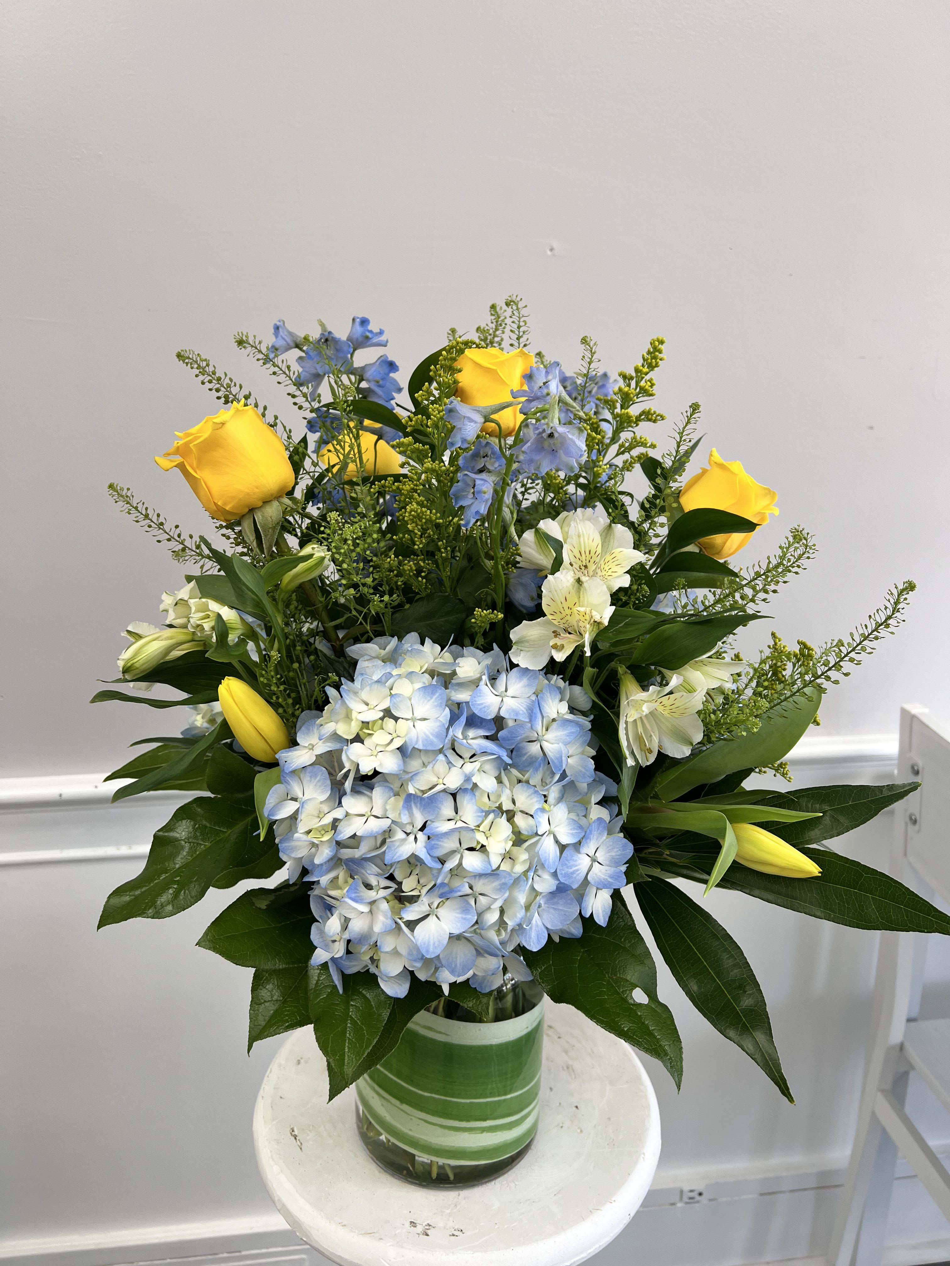 Mixed arrangement of yellow roses and tulips, blue hydrangea, and white blooms in a glass vase with green wrap