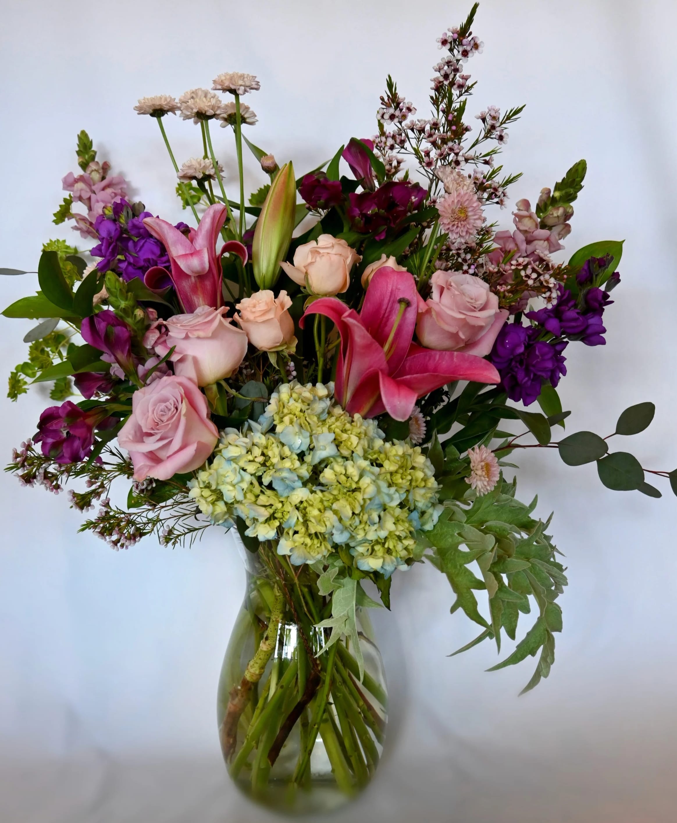 Empress Bloom: Flor de Emperatriz - Indulge in the grandeur of our towering garden-style arrangement, crafted to captivate the senses. Adorned with lush hydrangeas, graceful snapdragons, romantic roses, fragrant stock, lilies, spray roses, alstroemeria, dutch poms, and a touch of seasonal splendor, this magnificent display is presented in a crystal-clear Jordan vase.  While this Spring-themed masterpiece is a delightful choice for any occasion, it proudly stands tall to pay homage to Mother's Day or to express heartfelt gratitude to the caregivers and cherished individuals in your life.  DIMENSIONS: Approximately 20" Diameter x 25" Height // Sumérgete en la grandeza de nuestro imponente arreglo estilo jardín, meticulosamente elaborado para cautivar los sentidos. Adornado con exuberantes hortensias, elegantes boca de dragón, románticas rosas, fragantes clavellinas, exquisitos lirios, delicadas rosas en spray, vibrantes alstroemerias, encantadores pompones holandeses y un toque de esplendor estacional, esta magnífica exhibición se presenta en un jarrón de cristal claro estilo Jordan.  Aunque esta obra maestra temática de primavera es una elección encantadora para cualquier ocasión, se erige con orgullo para rendir homenaje al Día de la Madre o para expresar un sincero agradecimiento a los cuidadores y personas queridas en tu vida.  DIMENSIONES: Aproximadamente 20" de diámetro x 25" de altura