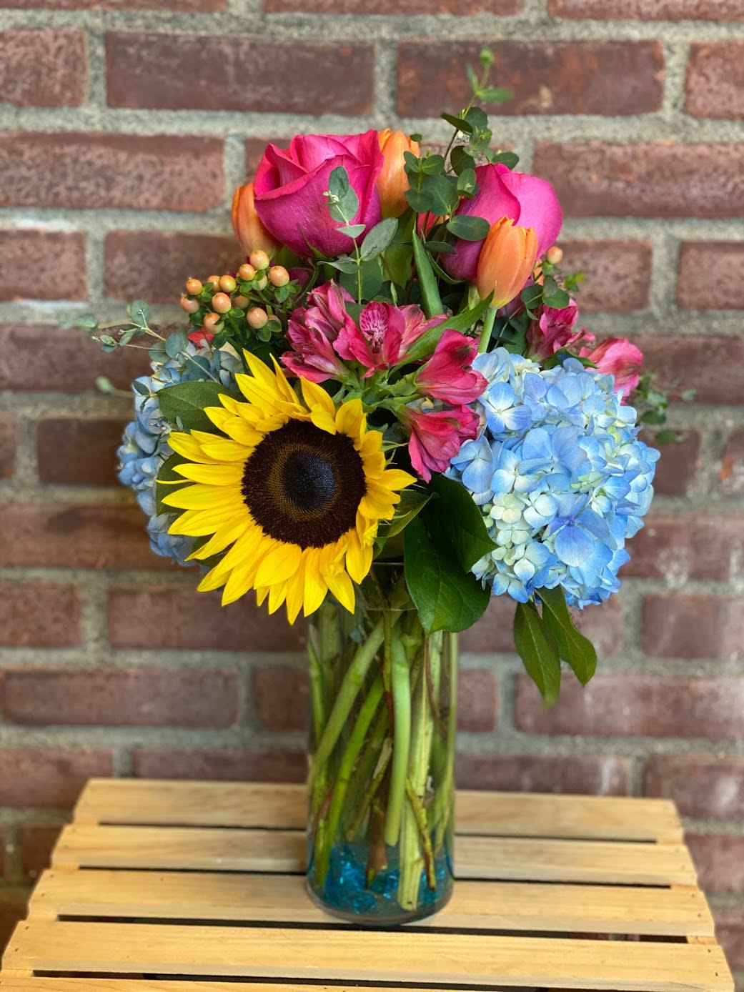 Bouquet of sunflowers, pink roses, blue hydrangeas, and orange tulips in a glass vase