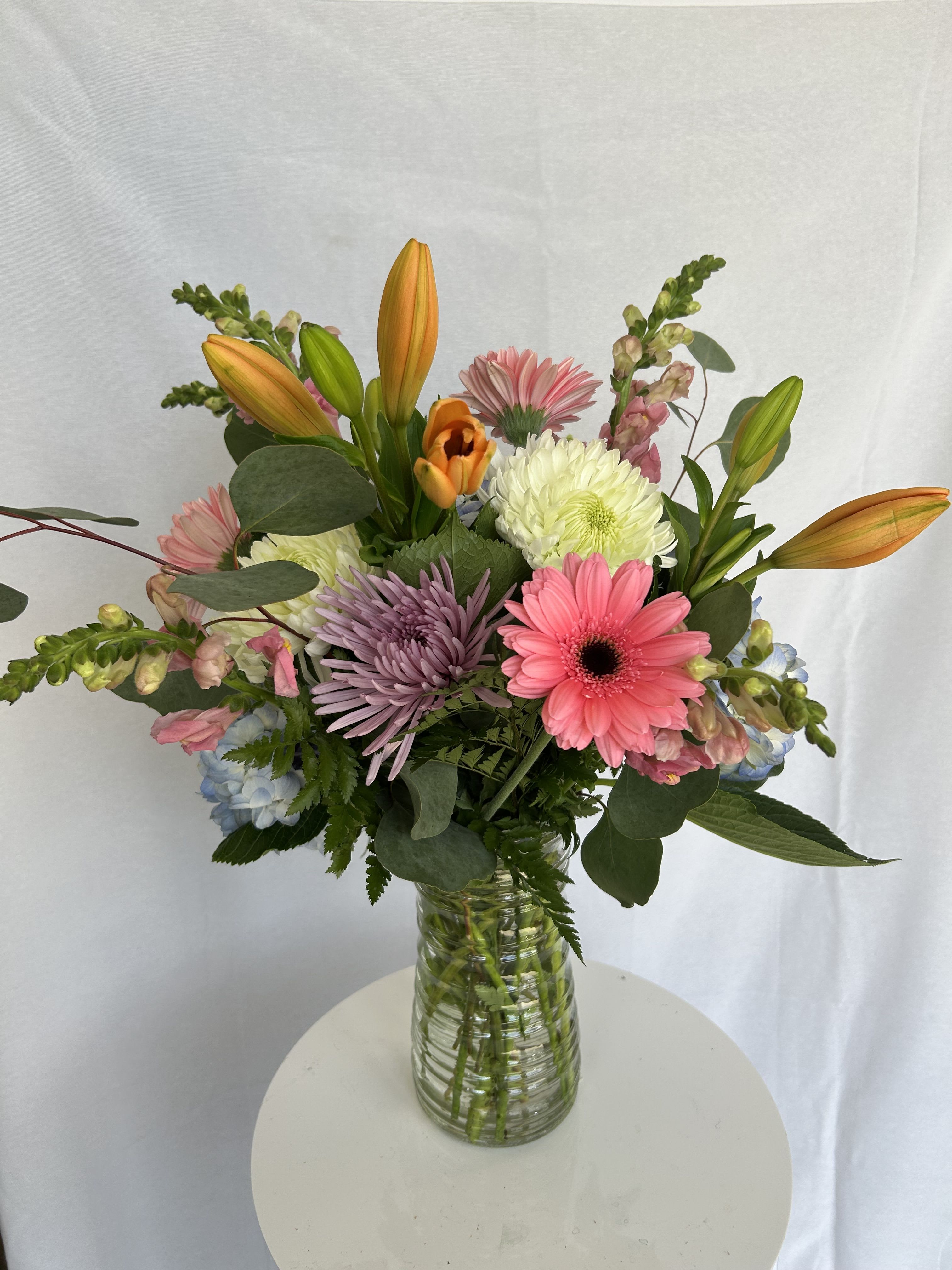 Daisy darling  - A vibrant, garden-inspired arrangement designed to feel fresh, joyful, and effortlessly elegant. This bouquet features a playful mix of textures and colors—soft blush and coral tones paired with creamy whites, pops of lavender, and hints of sky blue. Bold gerbera daisies and lush chrysanthemums create a full, eye-catching base, while striking orange lilies add height and a touch of drama. Delicate snapdragons and seasonal greenery weave throughout, giving the design a natural, airy movement. Arranged in a clear glass vase, this piece is perfect for brightening up any space—whether you're celebrating, sending love, or simply treating yourself to something beautiful.